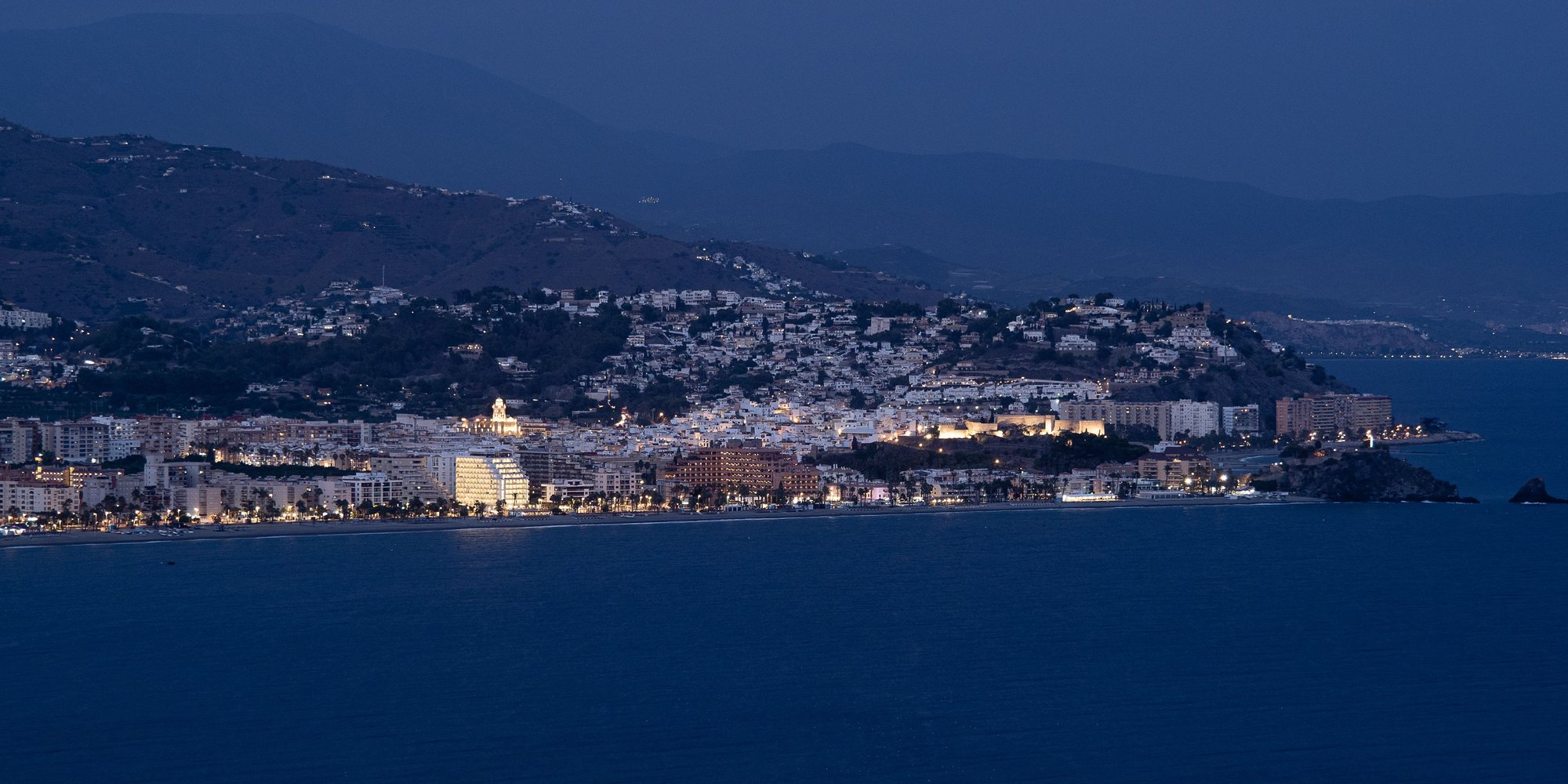 Almuñécar at dusk from the west, the floodlit castle on its hill above the white old town, the seafront lit along the bay and the Peñón del Santo rocks at the right