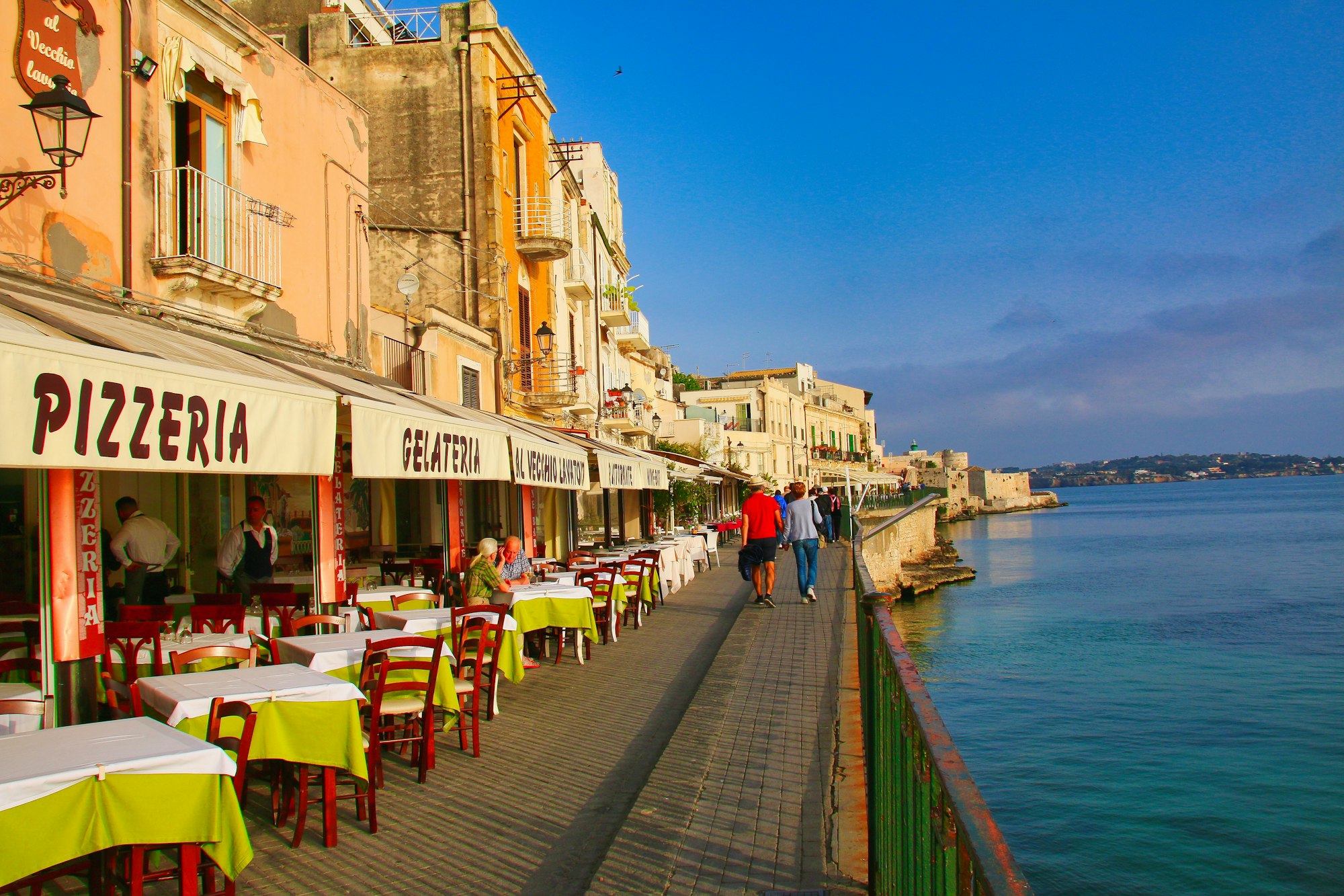 A seaside street of an Amalfi Coast town in the evening, tables and a trattoria sign beside the water