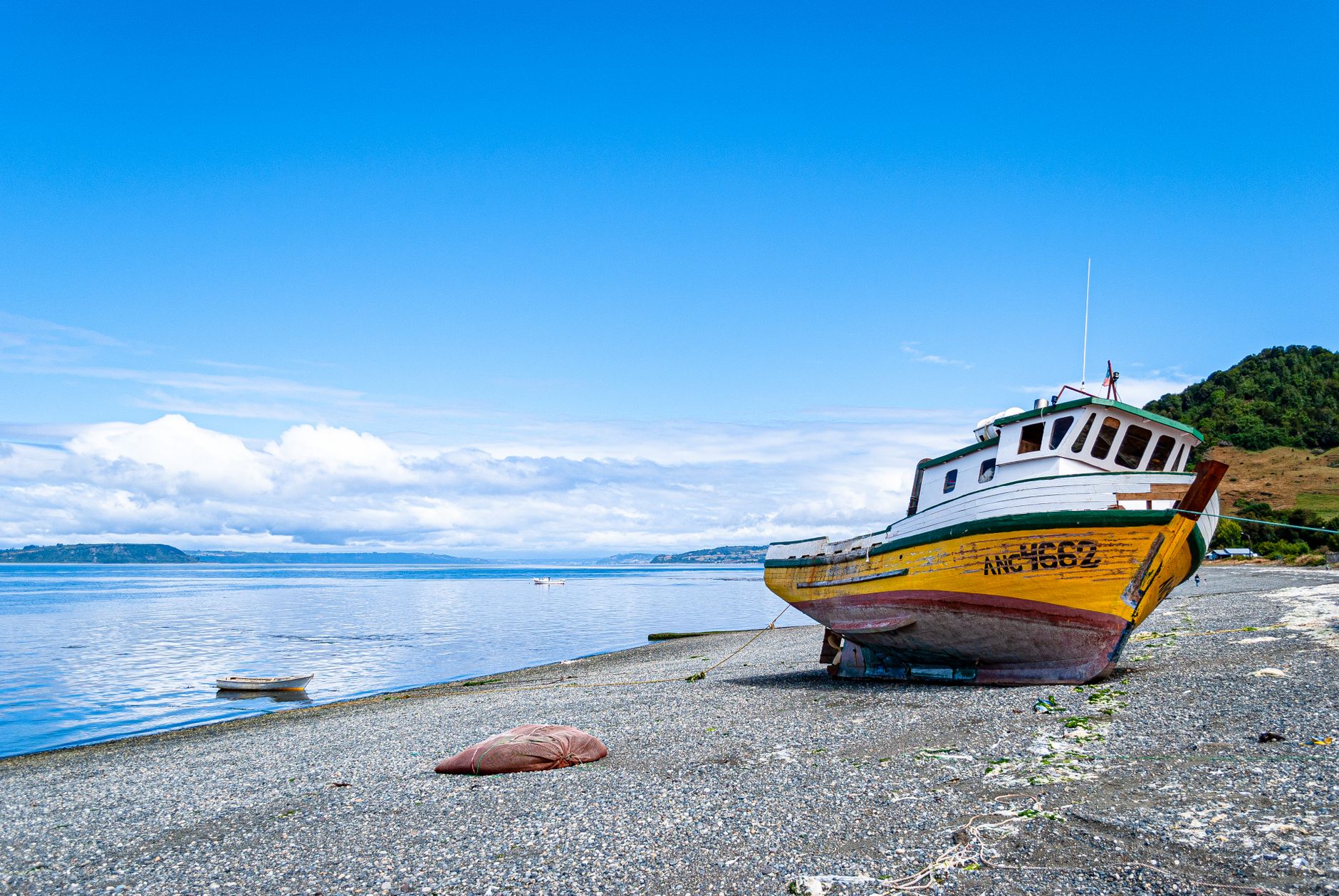 A yellow and white wooden fishing boat marked ANC 4662 standing on a shingle beach beside calm blue water