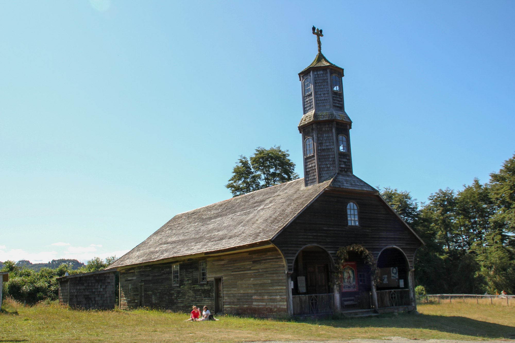 A weathered shingled wooden church with a tall tower and an arcaded porch on a grassy rise