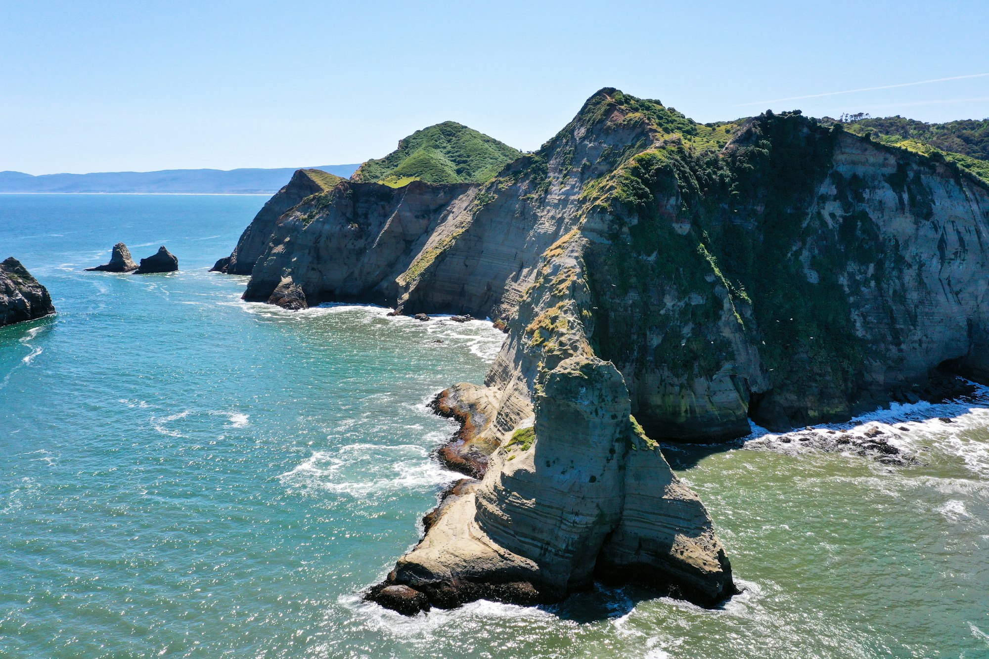 Green-topped cliffs and a pale rock stack above turquoise water on the Pacific coast