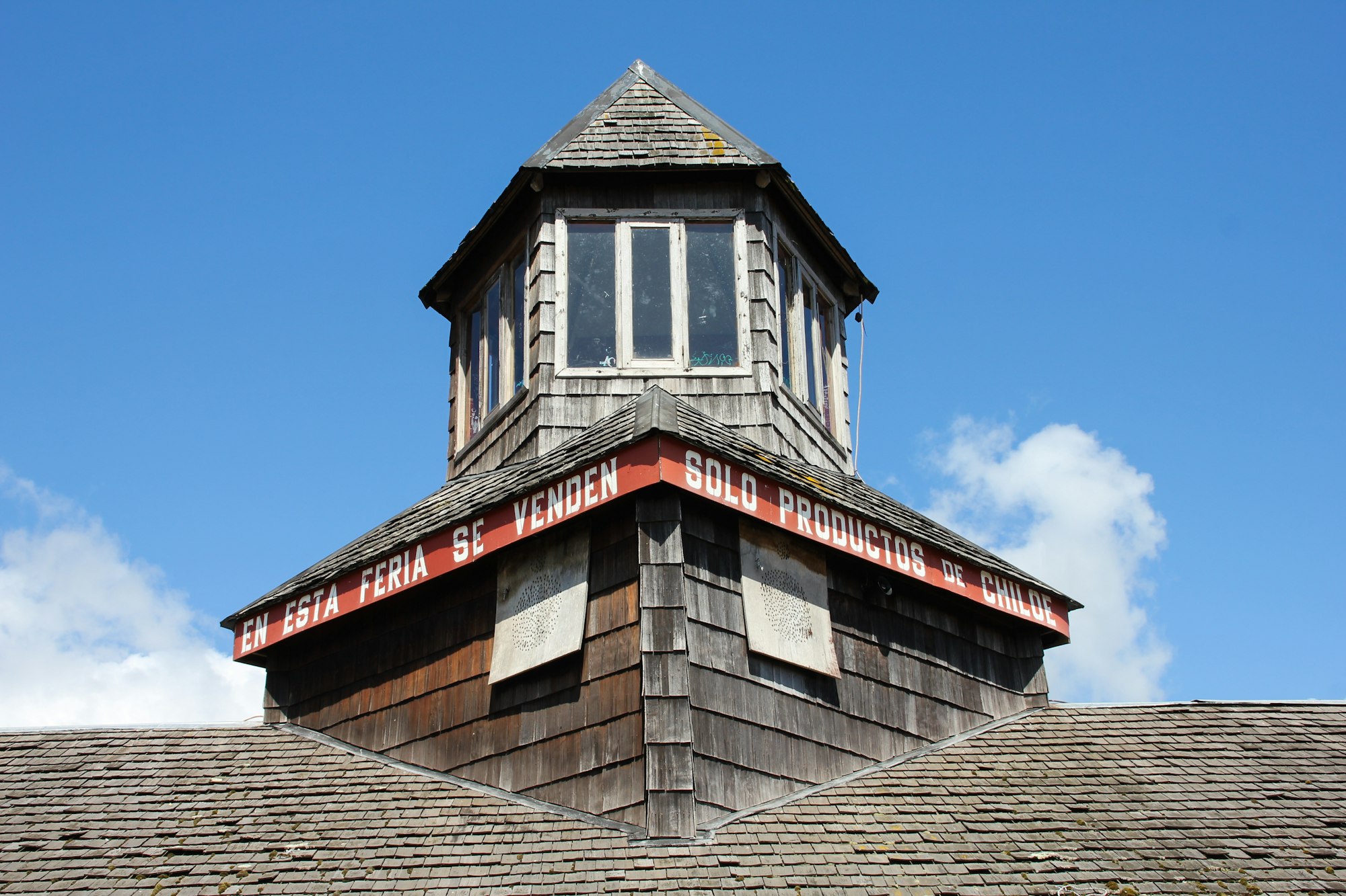 A wooden market tower clad in grey shingles with a red painted sign reading En esta feria se venden solo productos de Chiloé