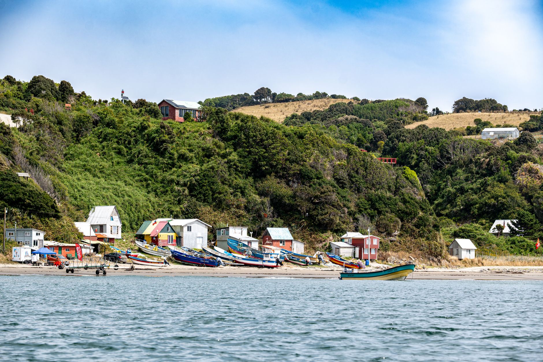 Brightly painted wooden houses and small fishing boats along a cove, with green hills and farmland behind