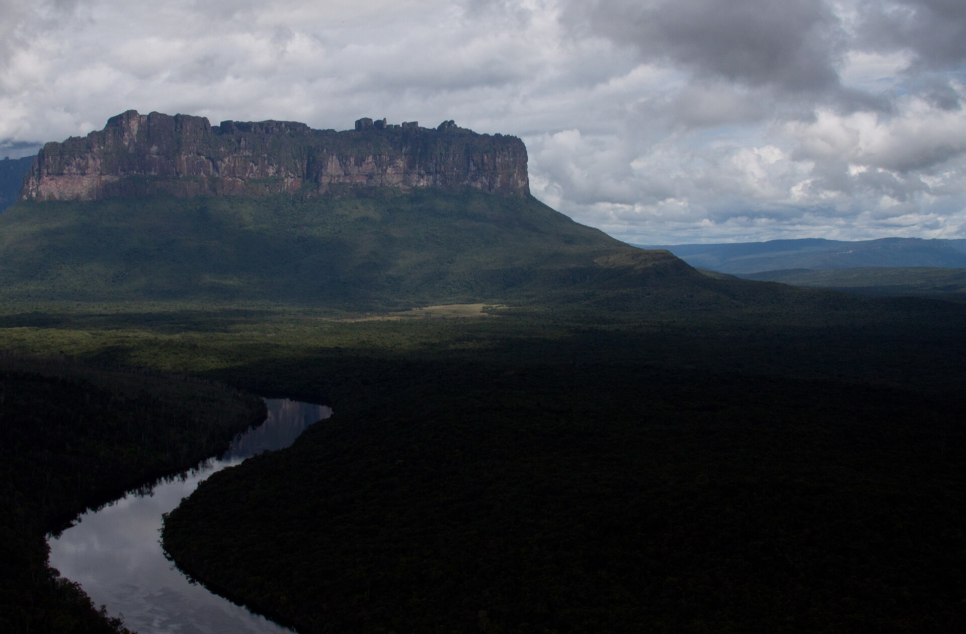 The flat summit and dark cliffs of Auyantepui rising above forest and a looping river under heavy cloud
