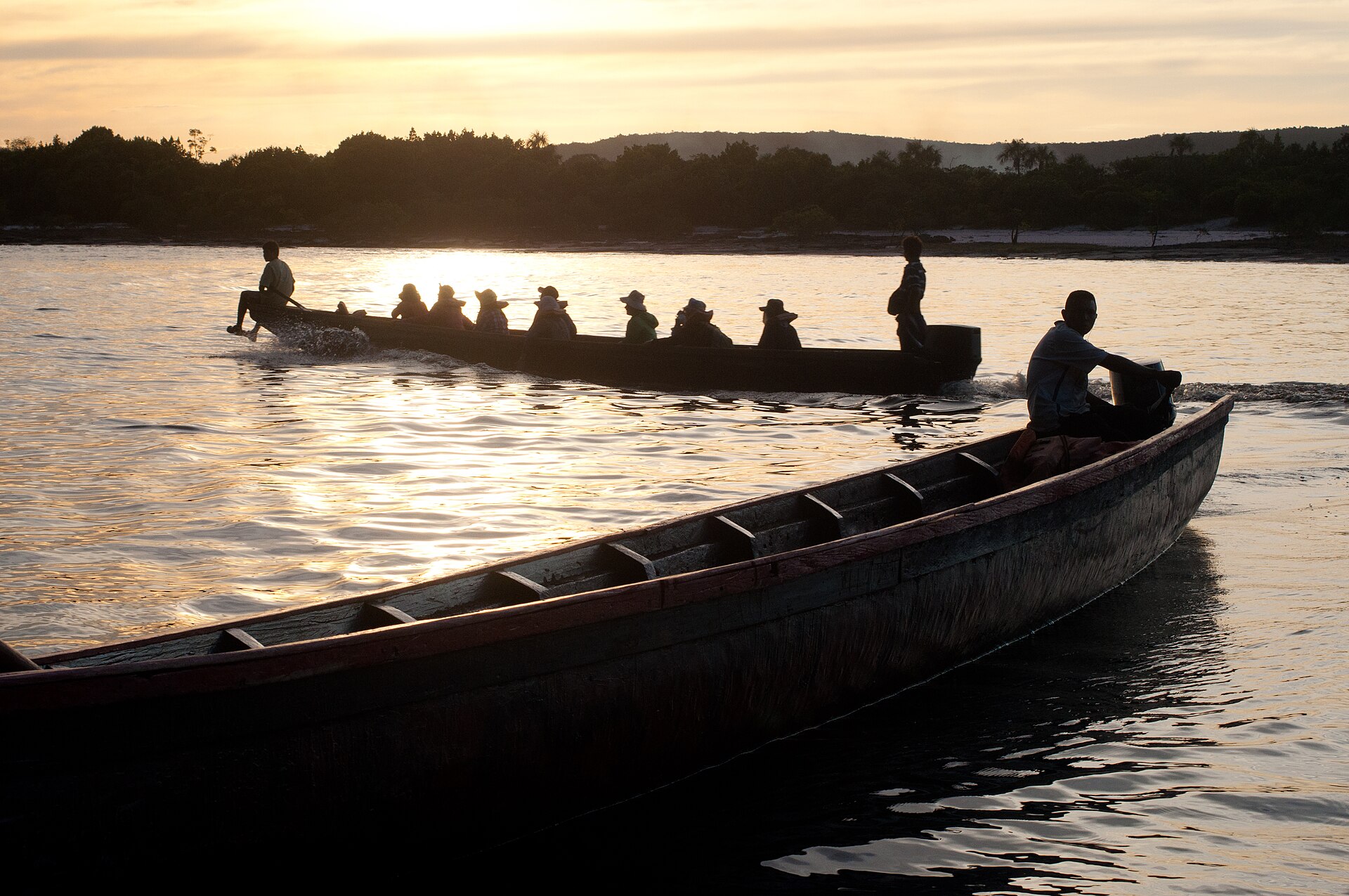 A long motorised dugout canoe carrying passengers across the Canaima lagoon at sunset