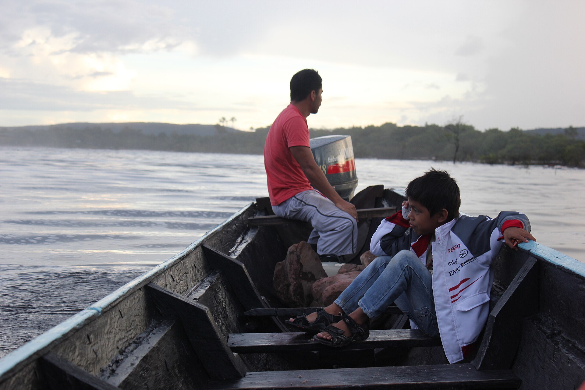 A Pemon boatman and a child sitting in a dugout on the water, the child taking a call on a mobile phone