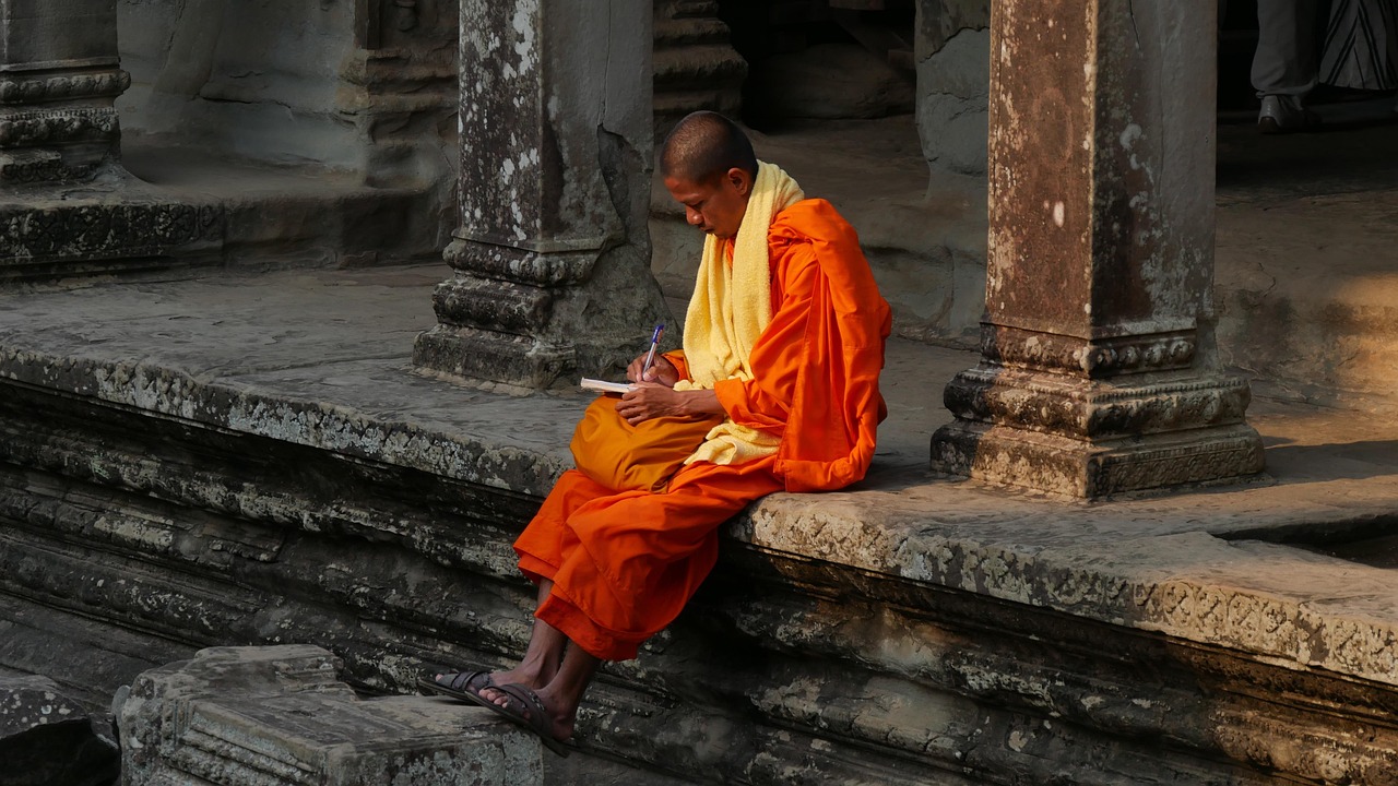 A monk in orange robes sitting reading in a stone colonnade at Angkor