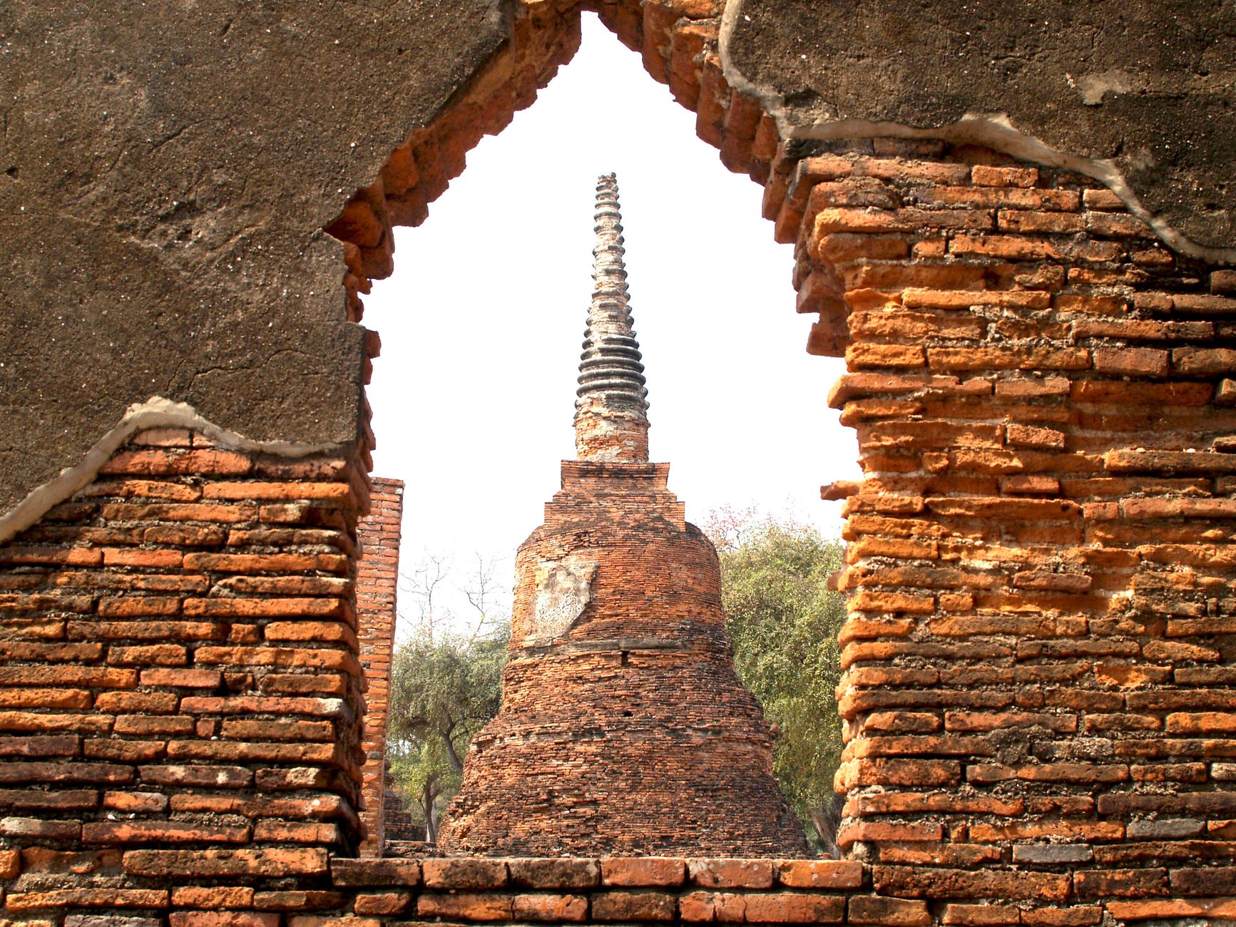 A view through a ruined brick window opening onto the spire of a chedi standing beyond