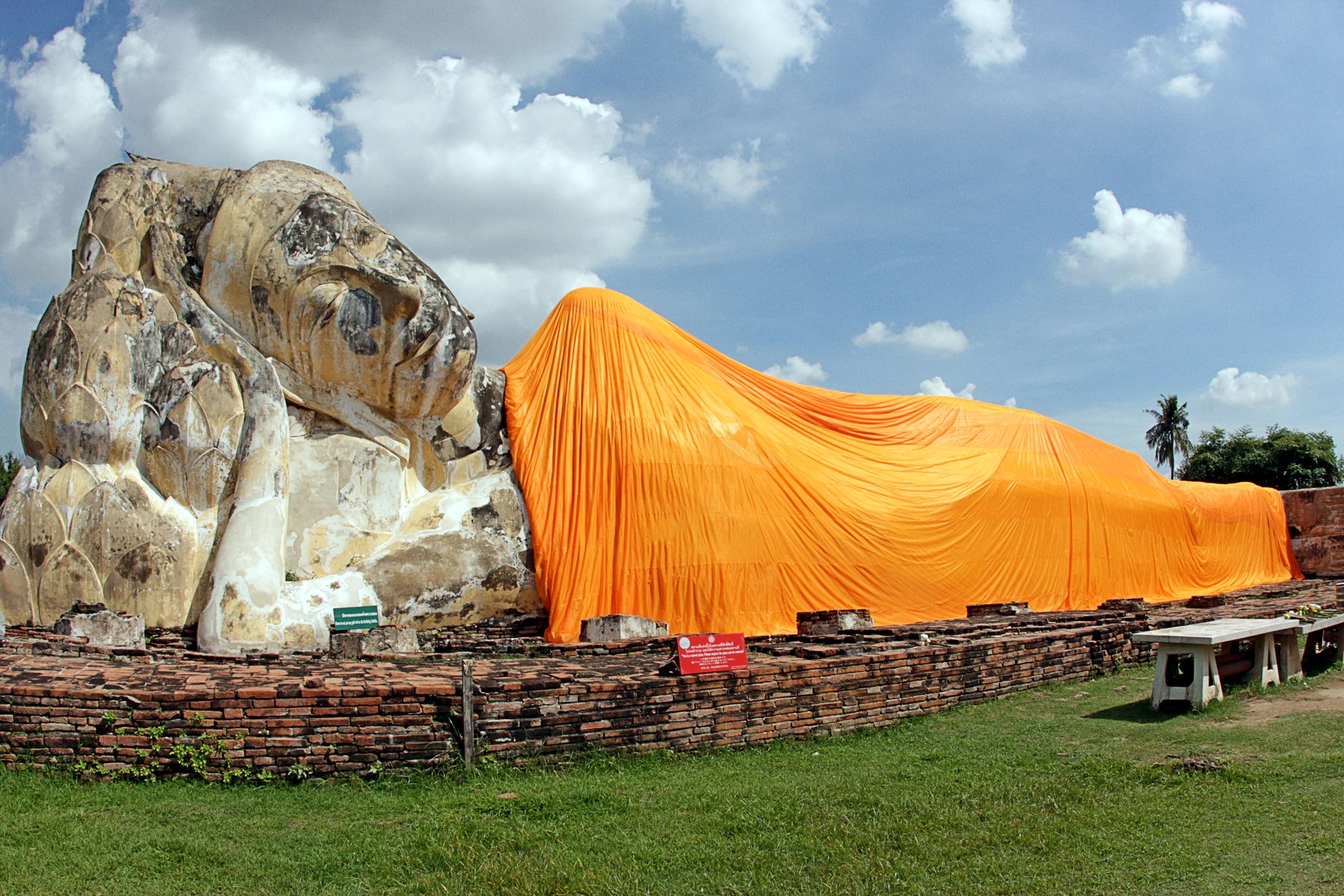 A long brick and stucco reclining Buddha draped in an orange robe lying in the open air with no roof above it