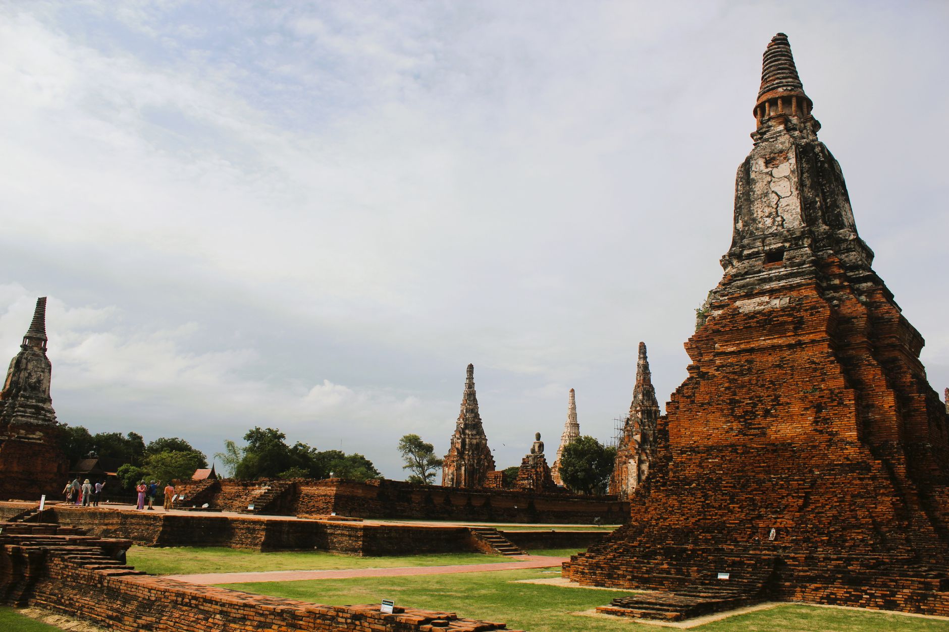 Ruined brick towers at Wat Chaiwatthanaram under an overcast sky, a corner meru filling the right of the frame, slender prangs and a seated Buddha behind, and visitors on the mown grass at left