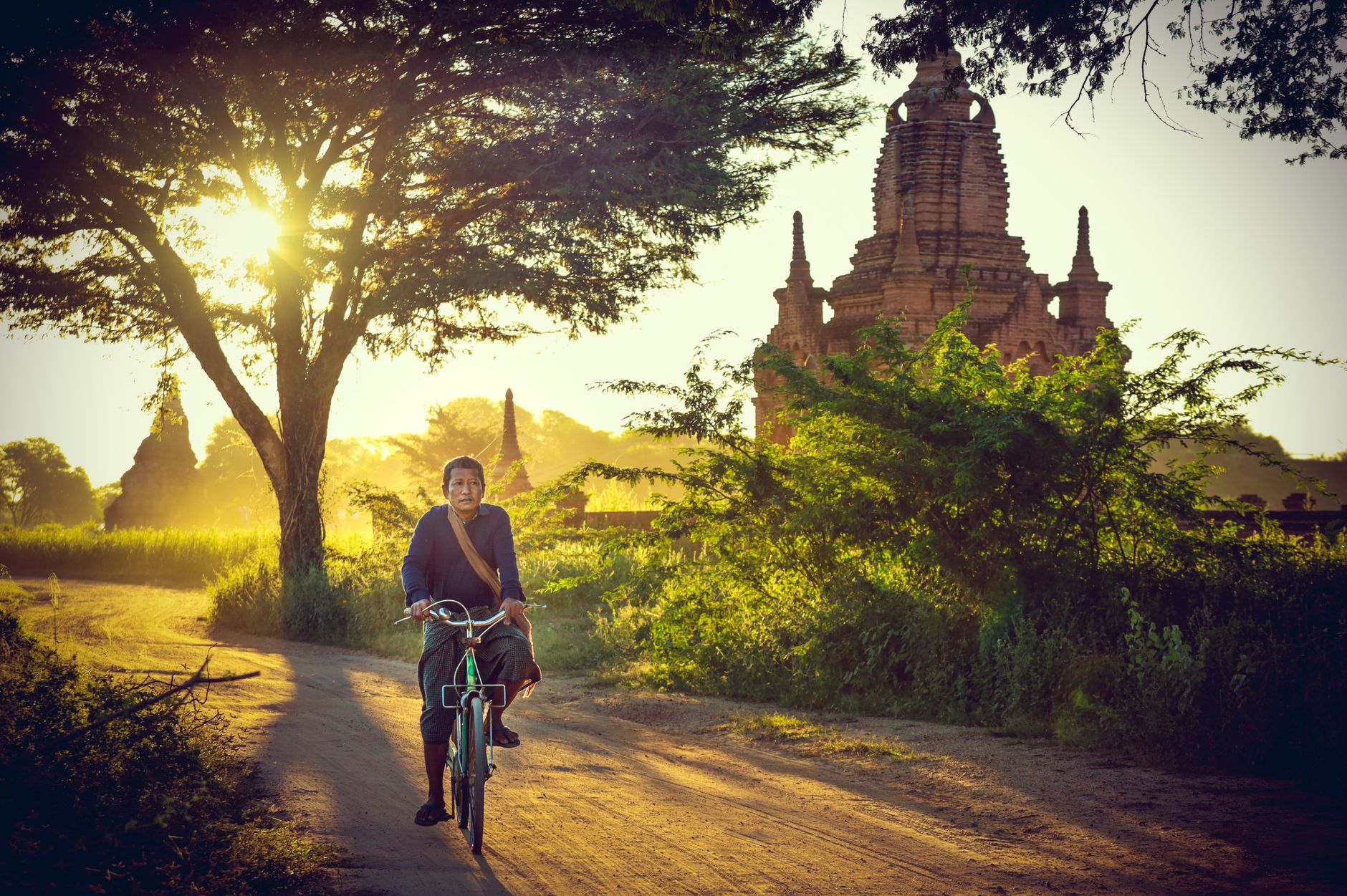 A man cycling along a sandy track past a brick temple in Bagan at golden hour