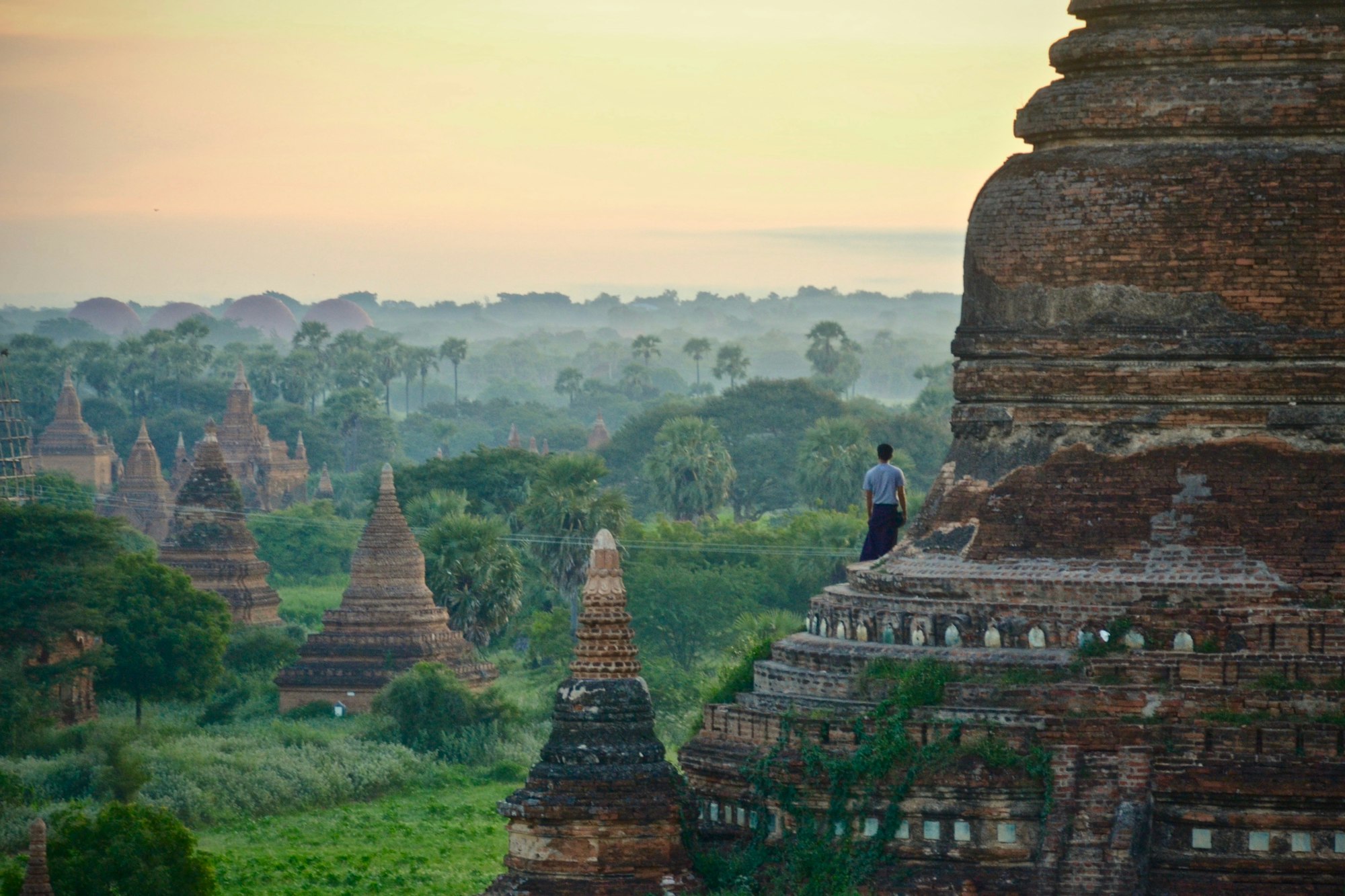Brick temple spires scattered across the Bagan plain in dawn haze, one figure standing on a temple terrace
