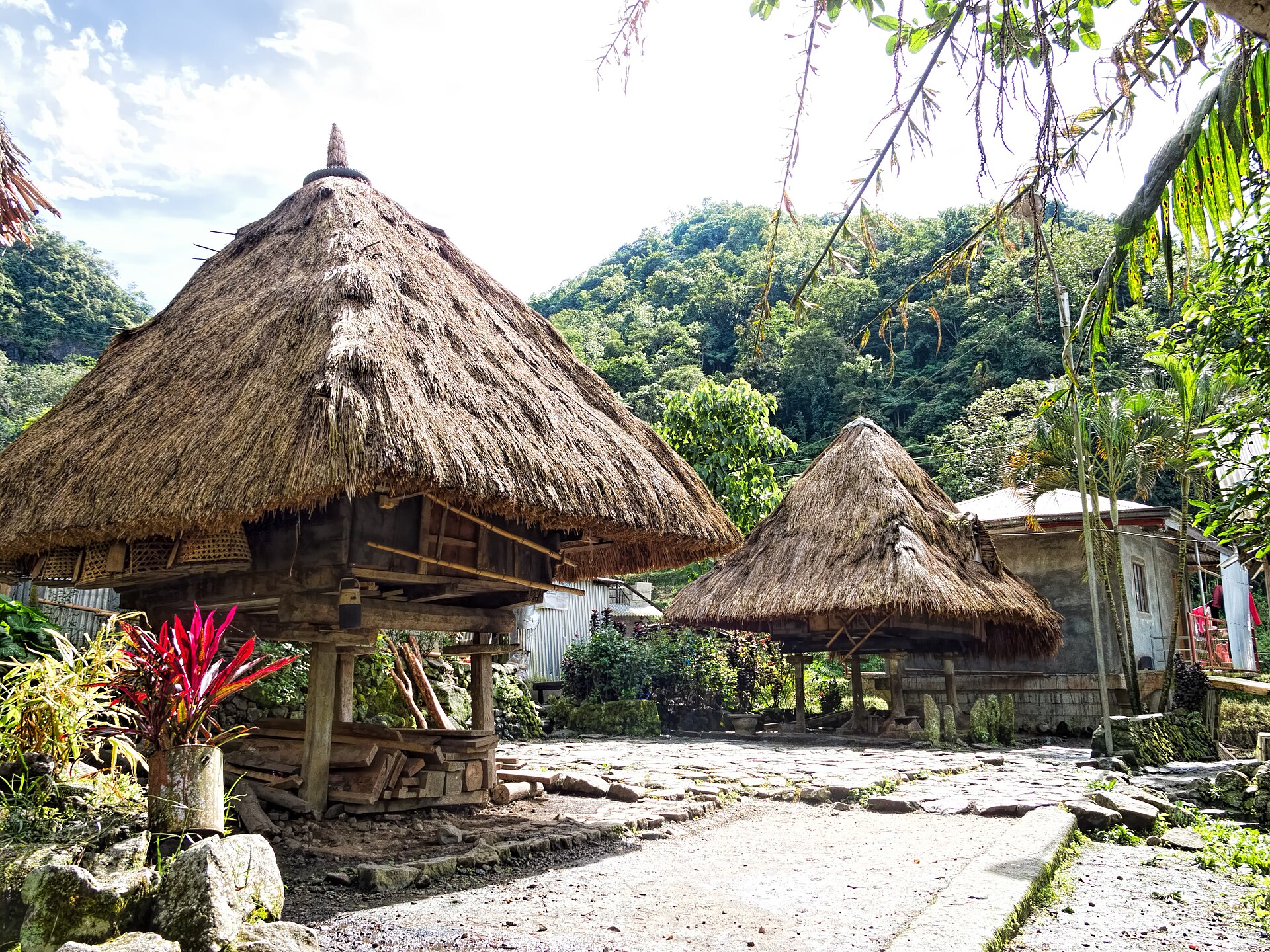 Two thatched pyramid-roofed Ifugao stilt structures on a stone-paved clearing at Bangaan