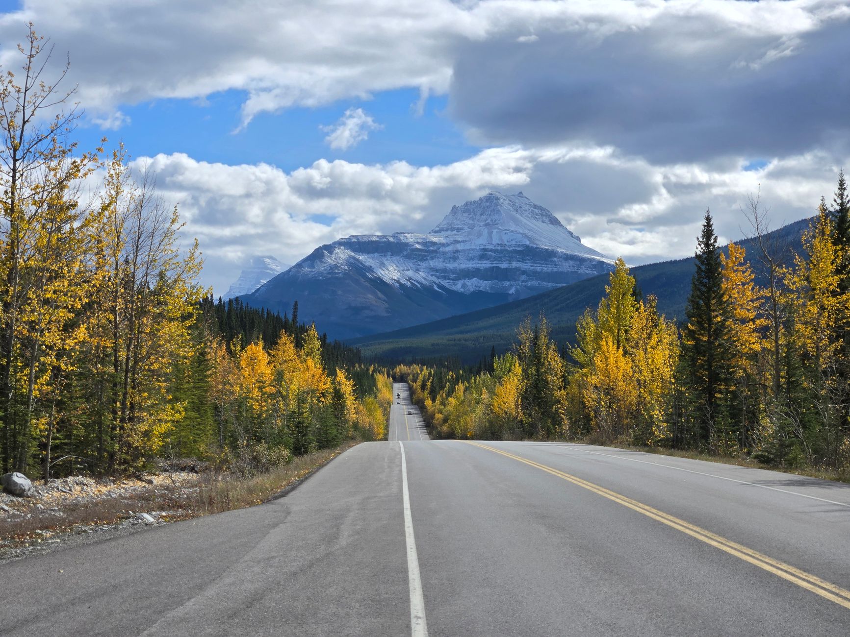 The two-lane Icefields Parkway running towards a large snow-streaked peak between autumn trees