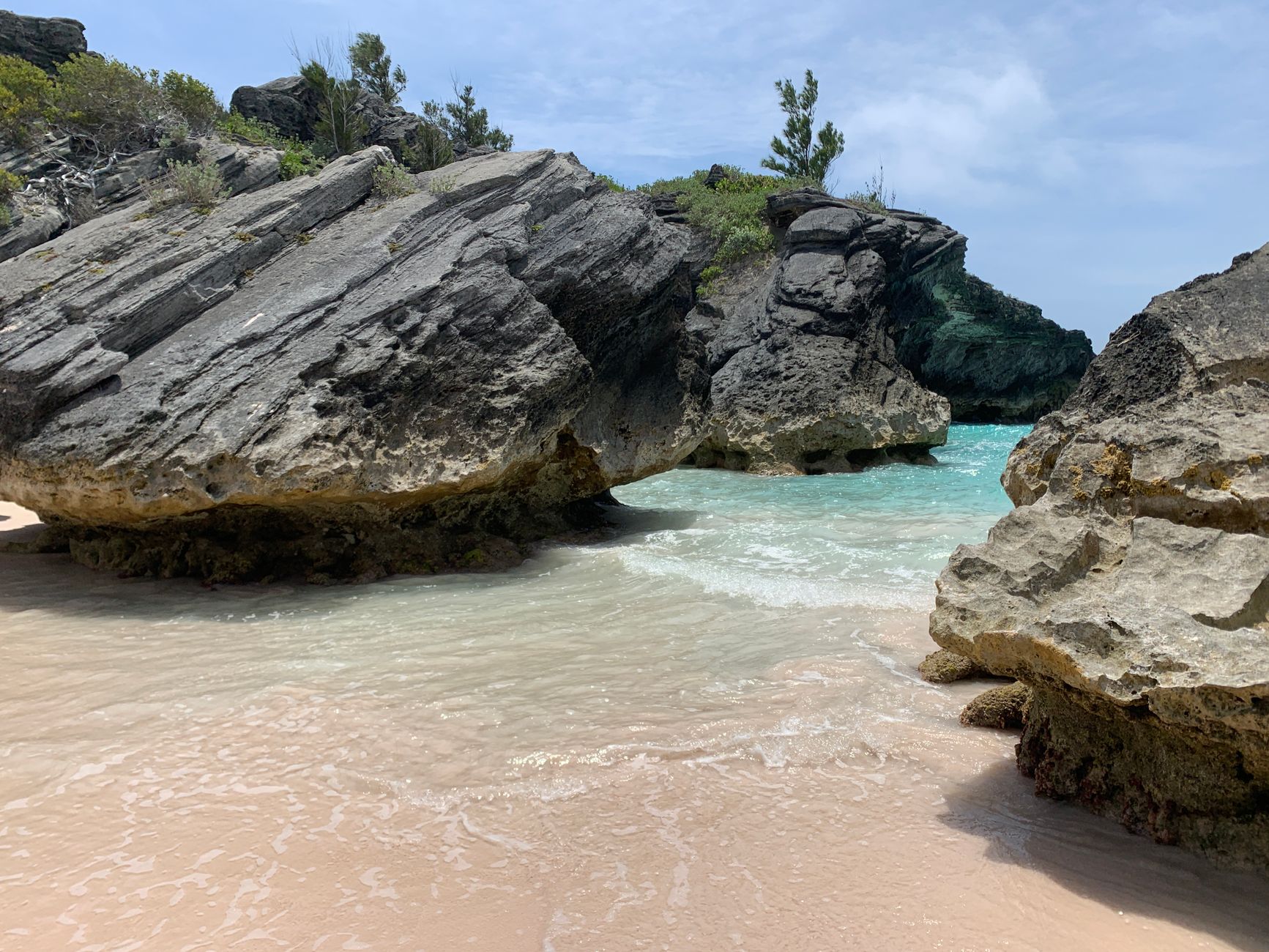Large weathered grey limestone boulders standing on pale pink tinged sand at the edge of clear turquoise water in a small Bermudian bay