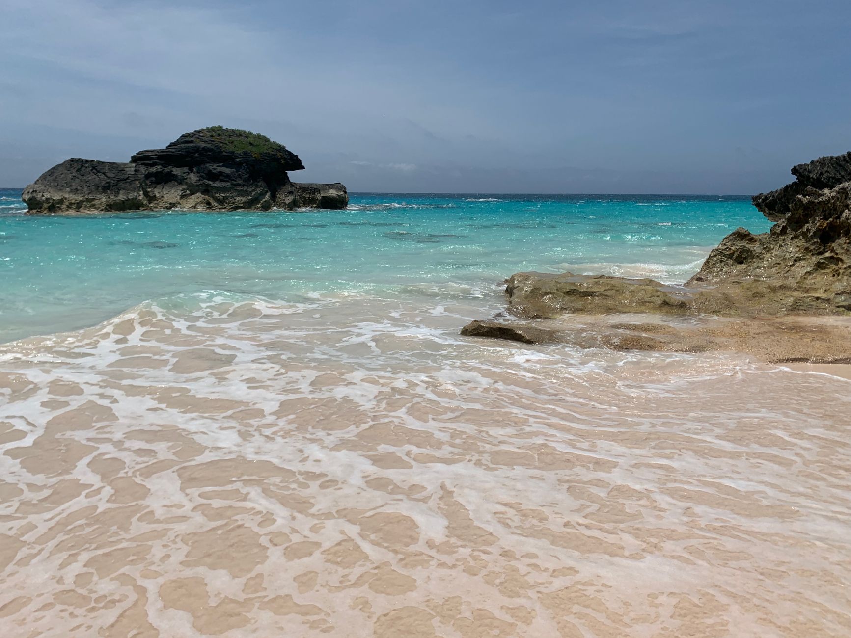 A sheltered Bermudian cove with pale sand, flat clear turquoise water and a dark limestone rock standing offshore