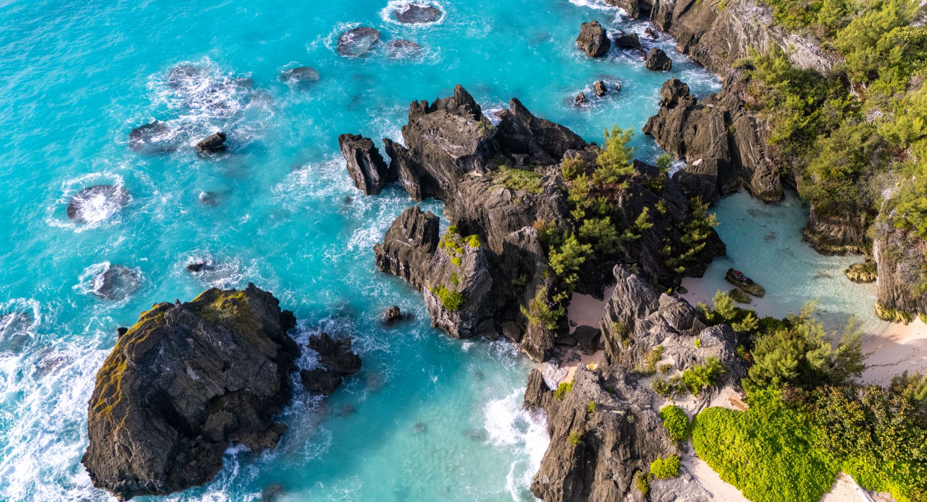 Aerial view of an eroded rocky headland with detached stacks and low vegetation lying in bright turquoise shallows beside a pale beach