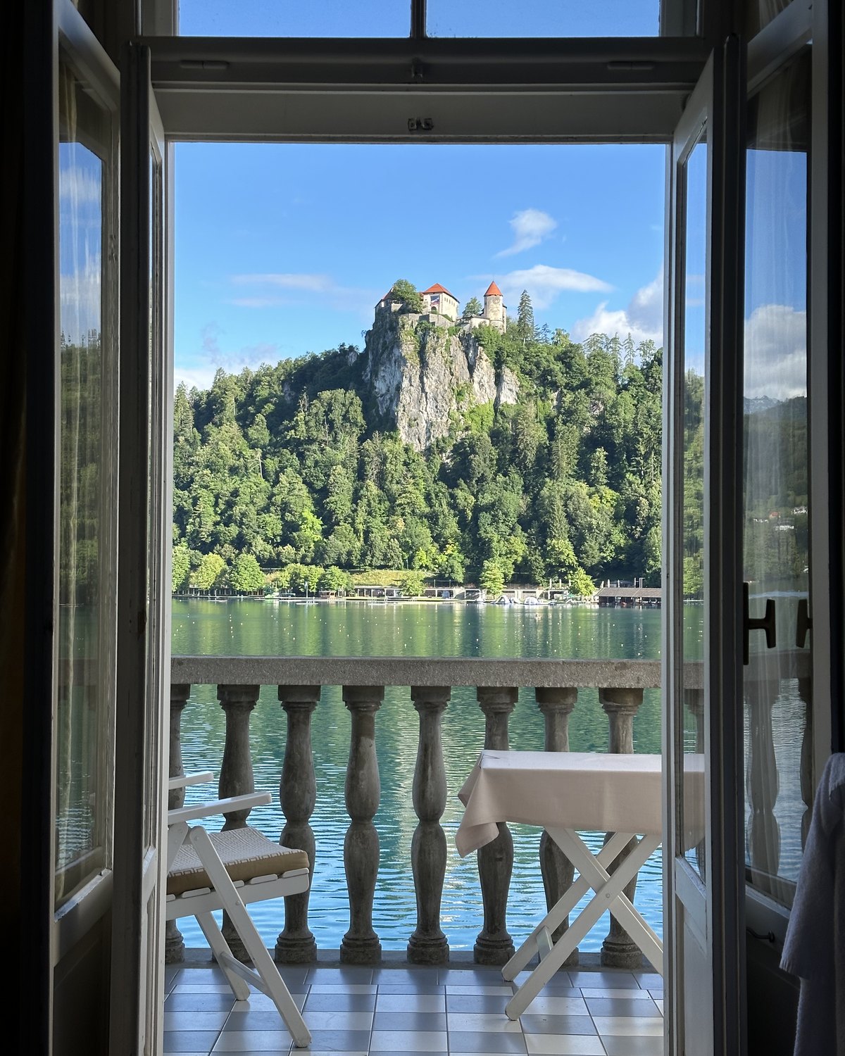 The view from a Grand Hotel Toplice room, open swing doors onto a stone balcony with a white chair and table, the lake and Bled castle beyond