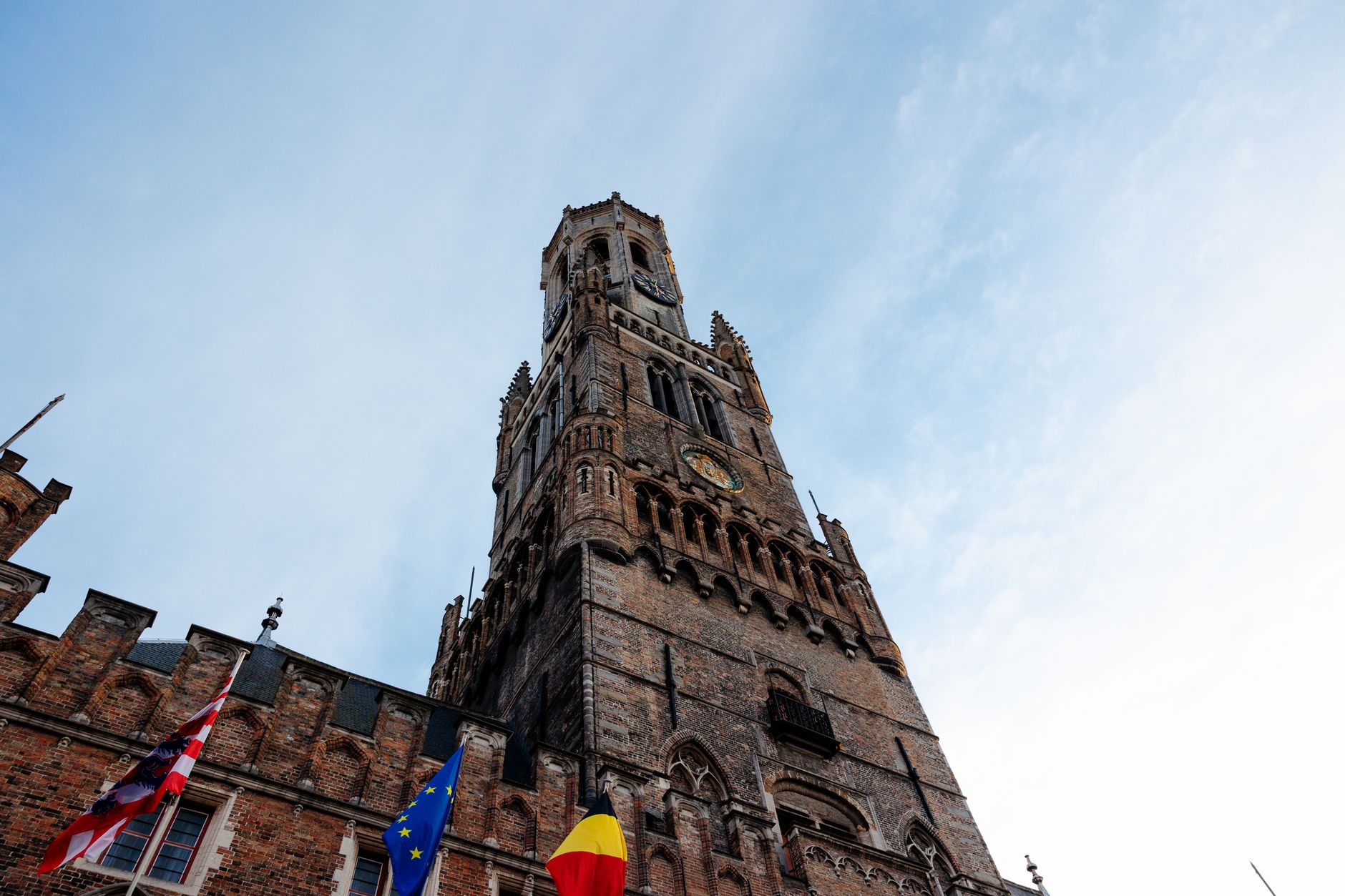 Low-angle view of the octagonal upper stage of the Belfry of Bruges against a clear blue sky, flags flying at its base