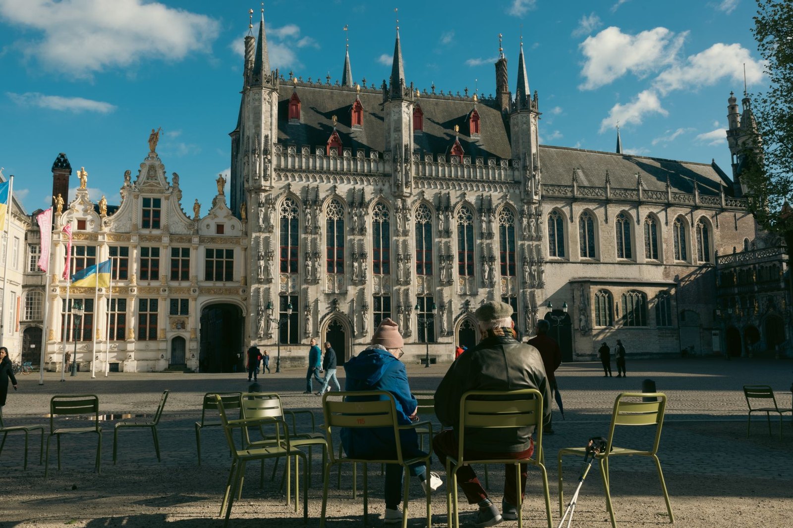 The Burg square in Bruges, the pale Gothic city hall with its turrets and statue niches in the centre, the gilded Renaissance front of the old civil registry to its left, and people sitting out on green metal chairs