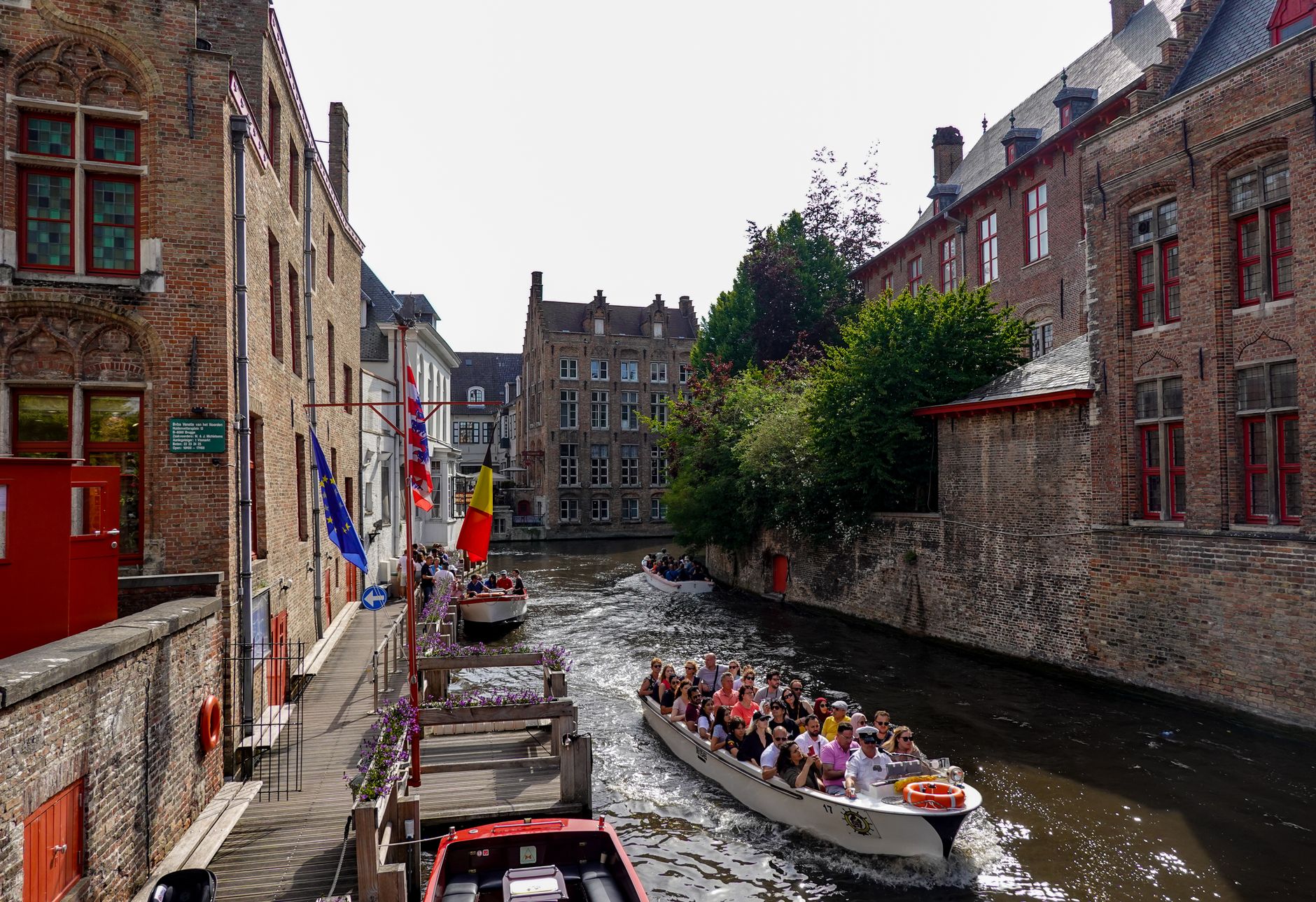 Three fully loaded open tour boats on a narrow Bruges canal, the Huidenvettersplein jetty sign on the quay behind