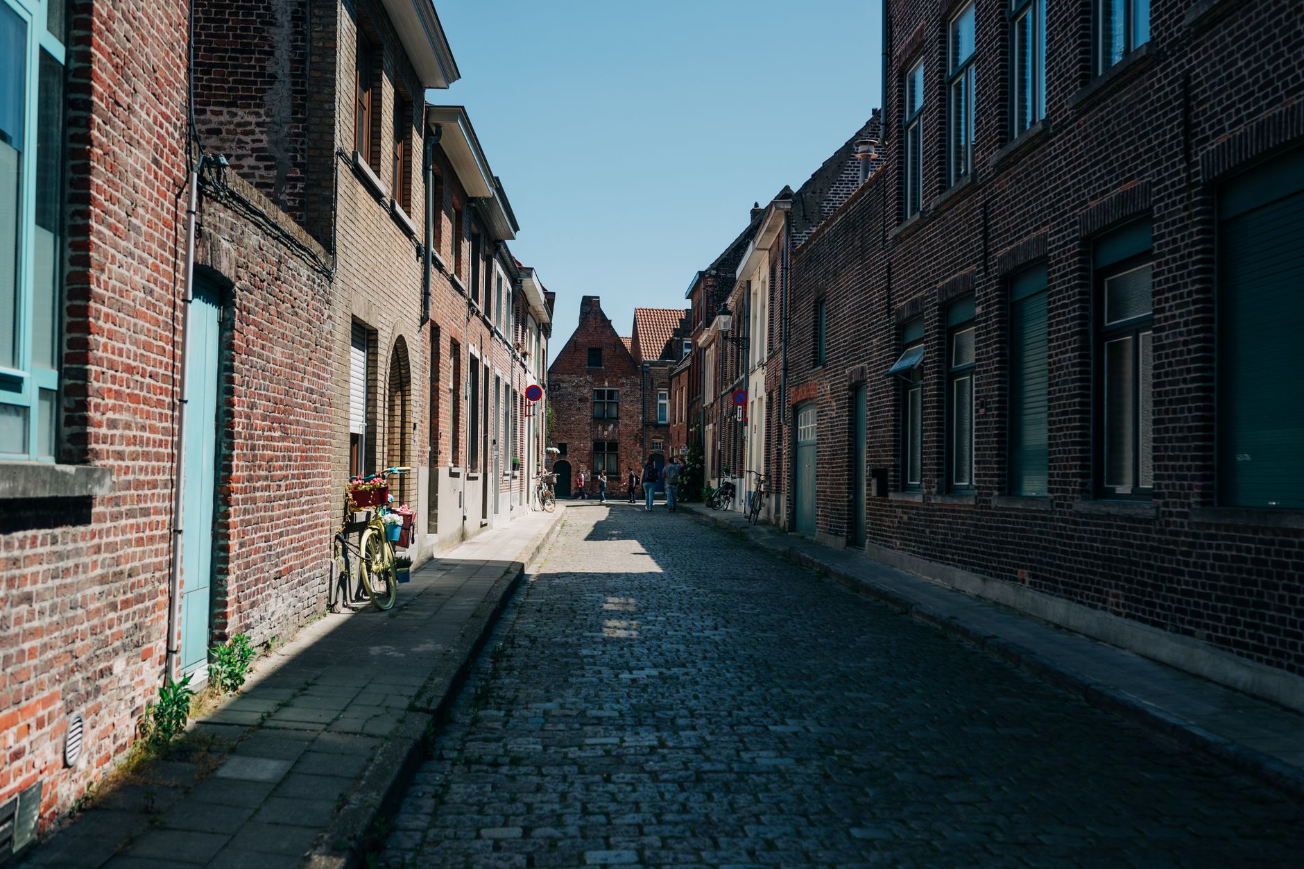 An empty cobbled lane of brick houses in Bruges closed by a stepped Flemish gable, one bicycle with a flower basket, hard midday sun