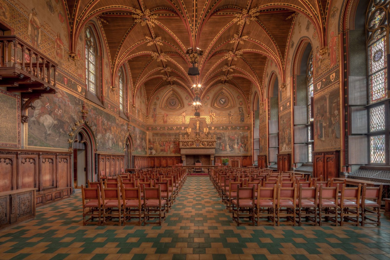 The Gothic Hall inside Bruges city hall, a painted timber vault with hanging bosses, murals along the walls and rows of red chairs on a tiled floor