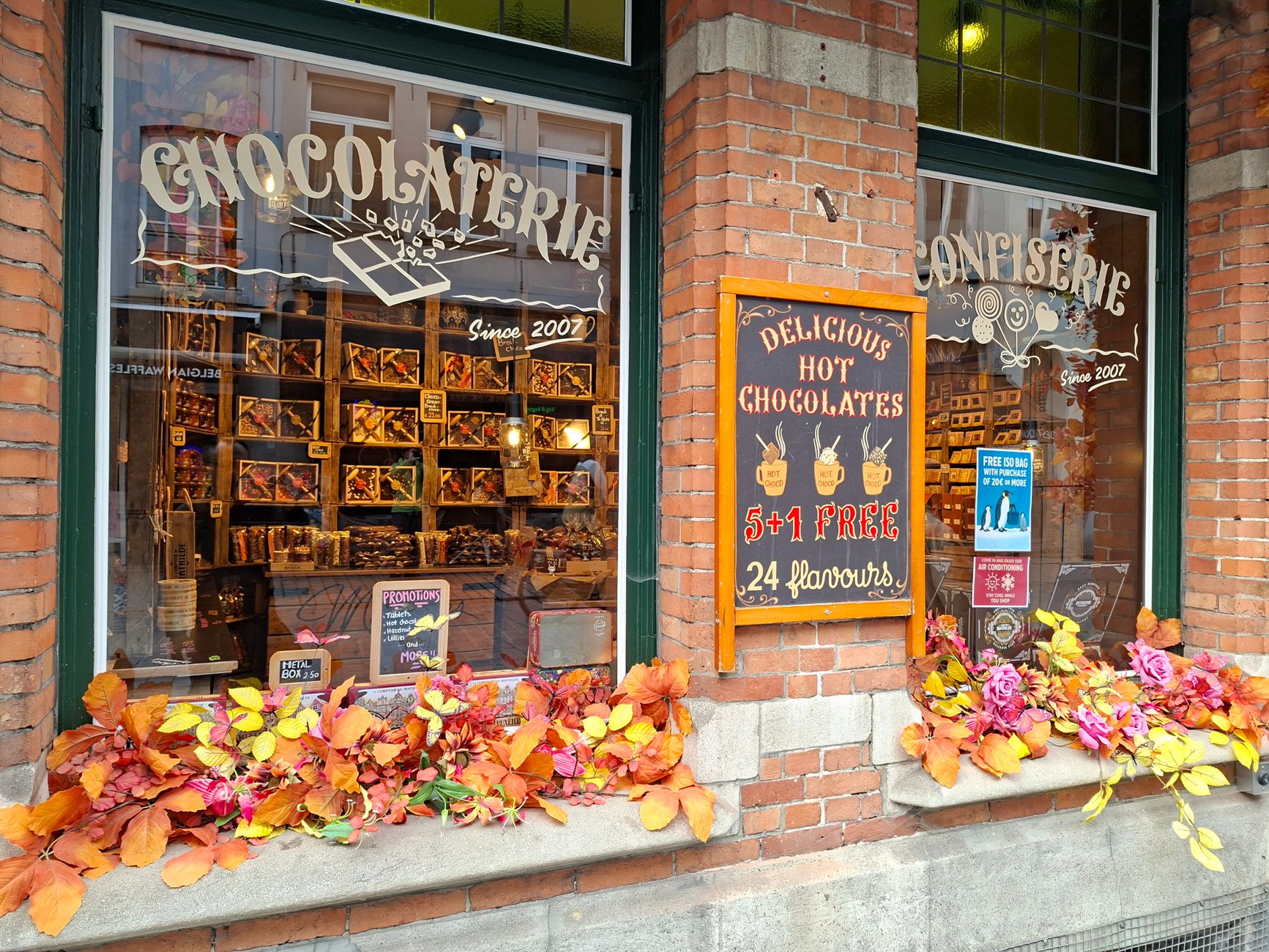 A bright chocolate shopfront in Bruges with dark green frames, CHOCOLATERIE and CONFISERIE painted on the glass, an autumn window box, and hot chocolate advertised on a chalkboard against the brick pier