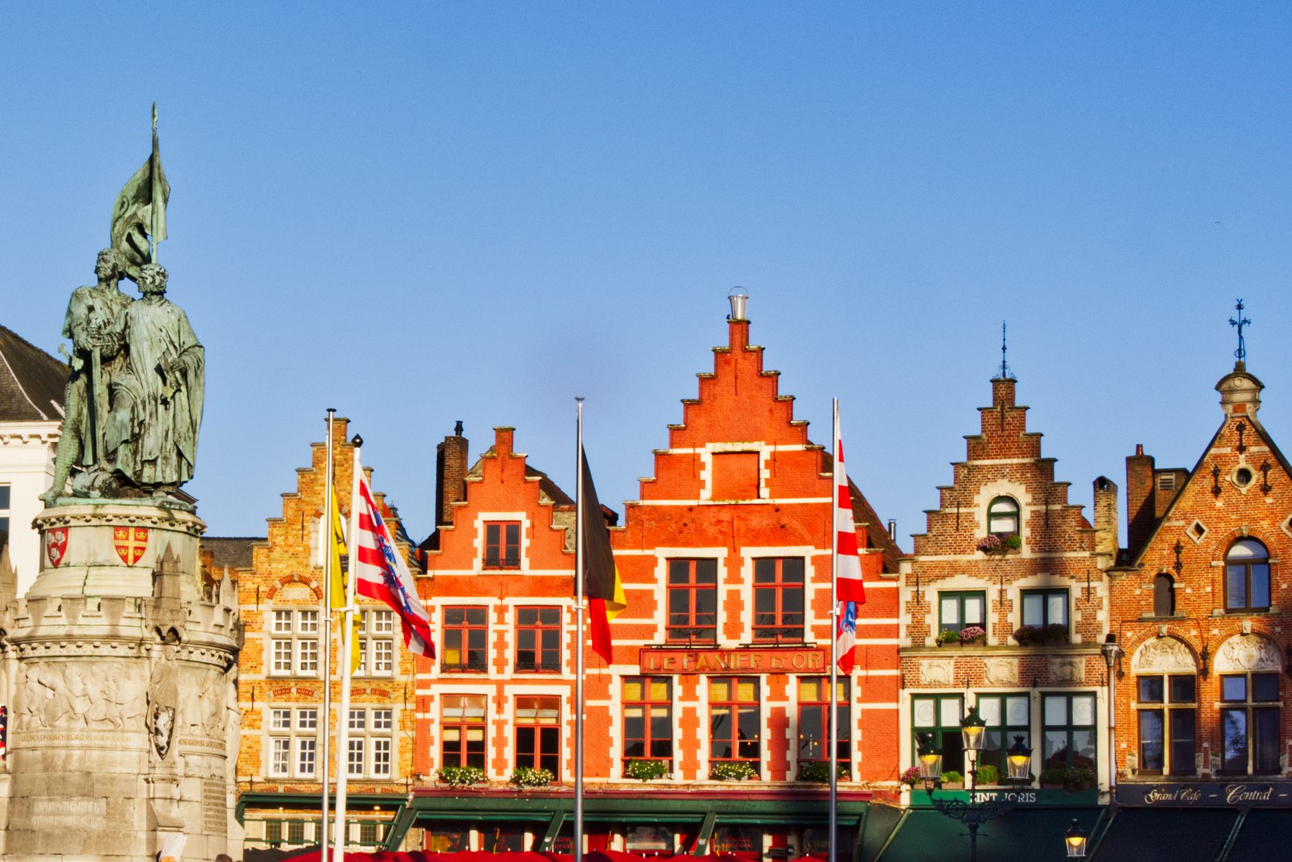 Stepped and bell-gabled guild houses in red, ochre and gold on the Markt in Bruges, flags flying, the Breydel and De Coninck statue at the left