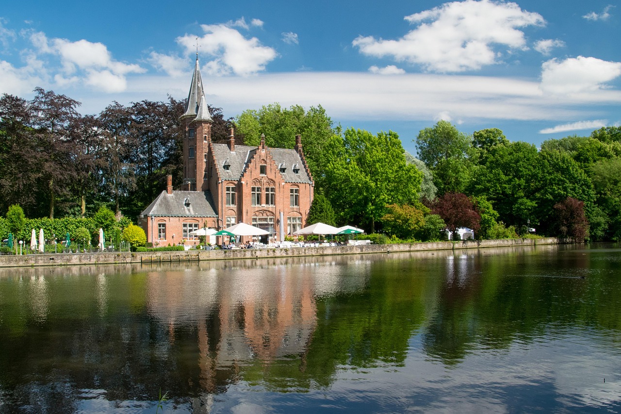 The red brick castle-style building with a slate spire on the bank of the Minnewater in Bruges, reflected in the lake under summer cloud