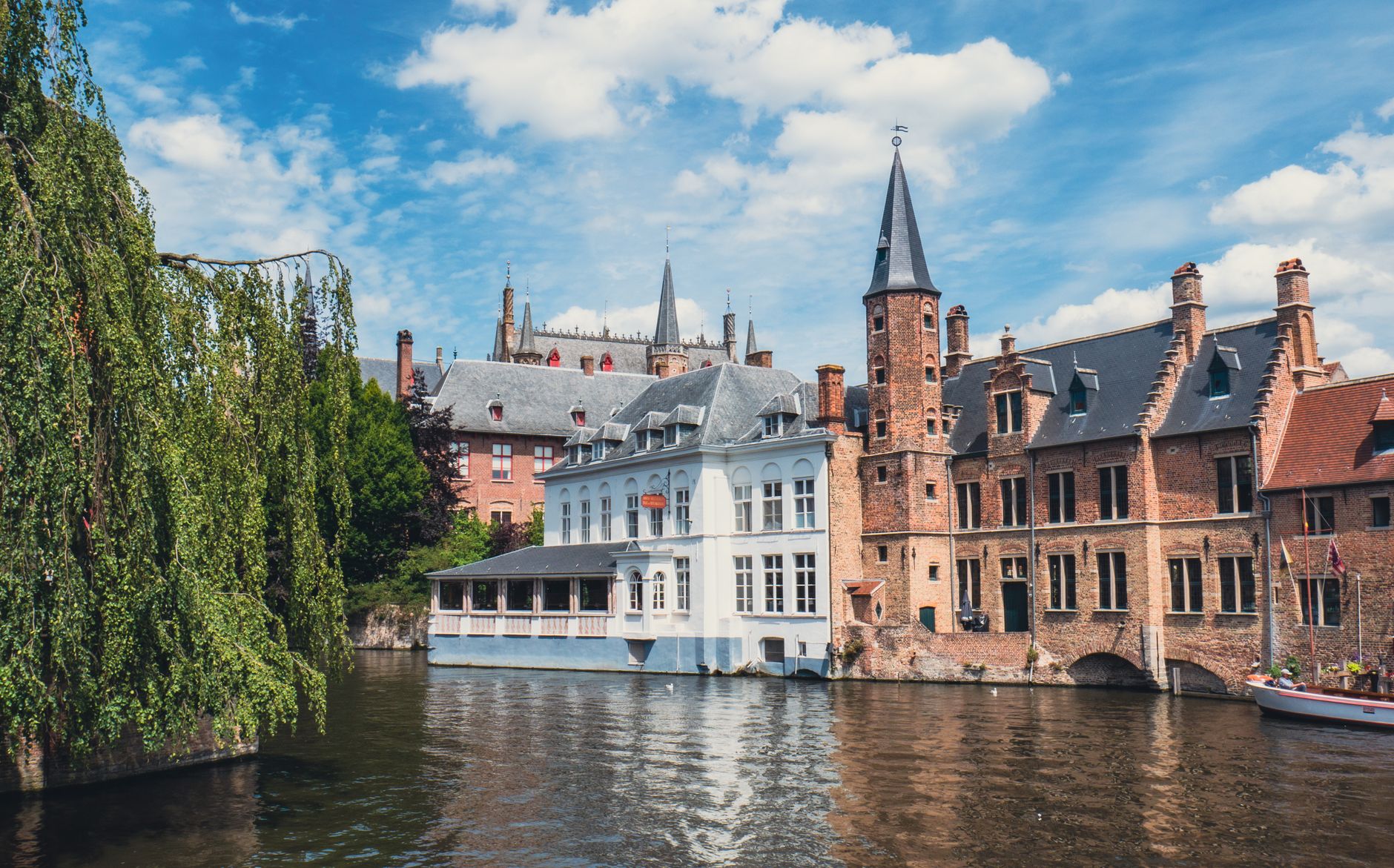 A Bruges canal frontage of stepped gables and a white rendered building with a glazed veranda over the water, willow leaning in from the left