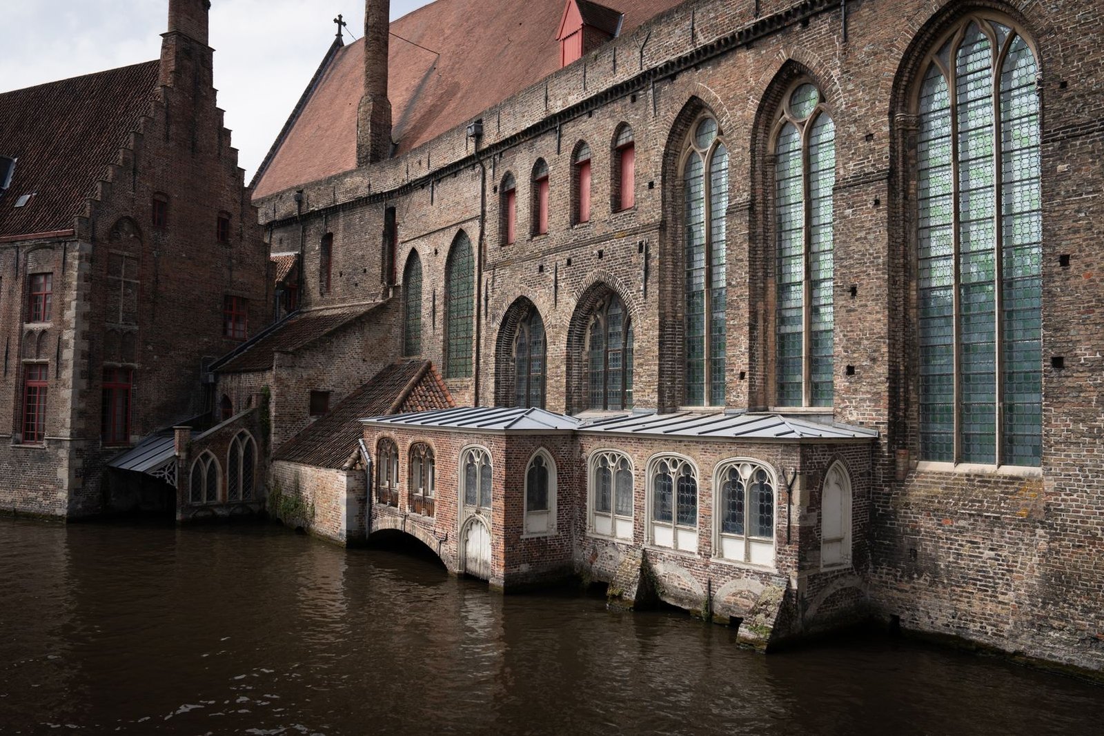 The long brick ward block and church of Sint-Janshospitaal in Bruges rising straight out of the canal, with tall pointed windows and a glazed lean-to built out over the water