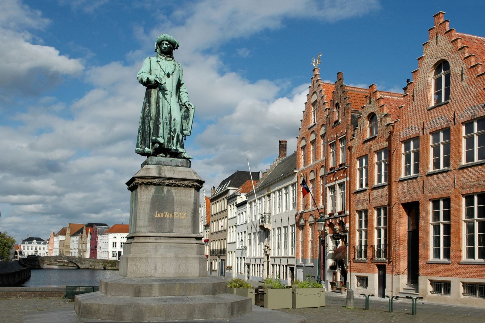 The bronze statue of Jan van Eyck on its plinth at Jan van Eyckplein in Bruges, gabled brick houses along the quay behind and the canal running off to the left