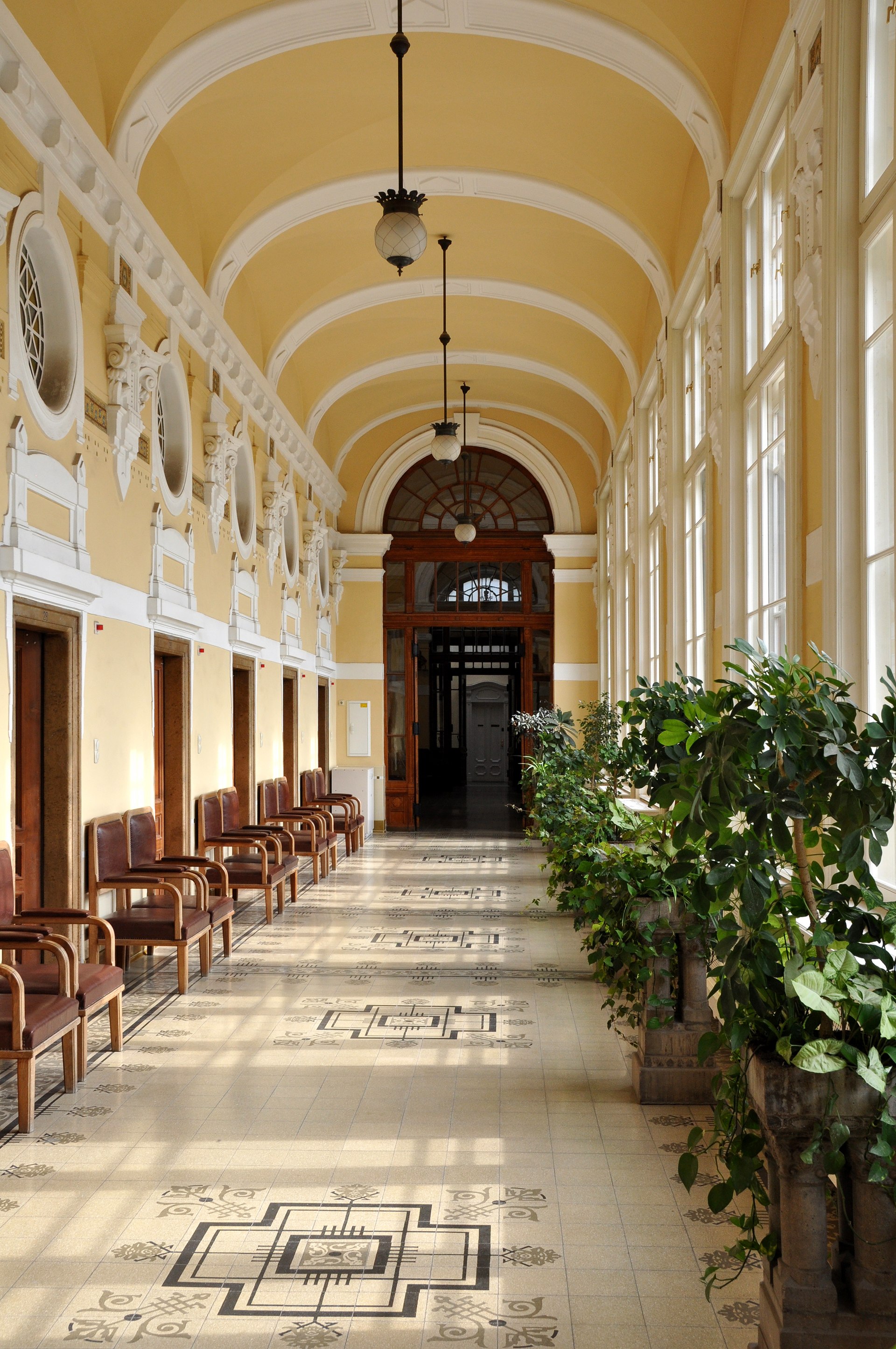 A long corridor inside the main building of the Szechenyi bath, with wooden chairs, potted plants and patterned floor tiling