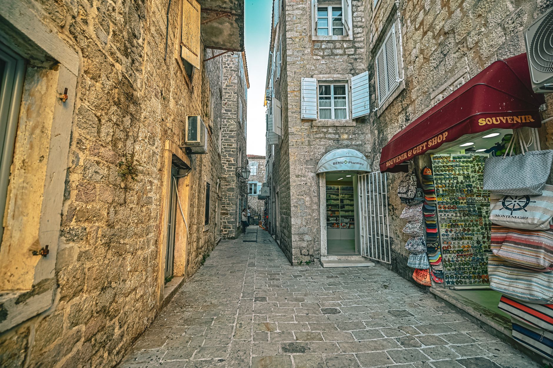 A narrow stone lane in Budva's old town with a souvenir shop awning on the right and a pharmacy doorway, empty flagstones underfoot
