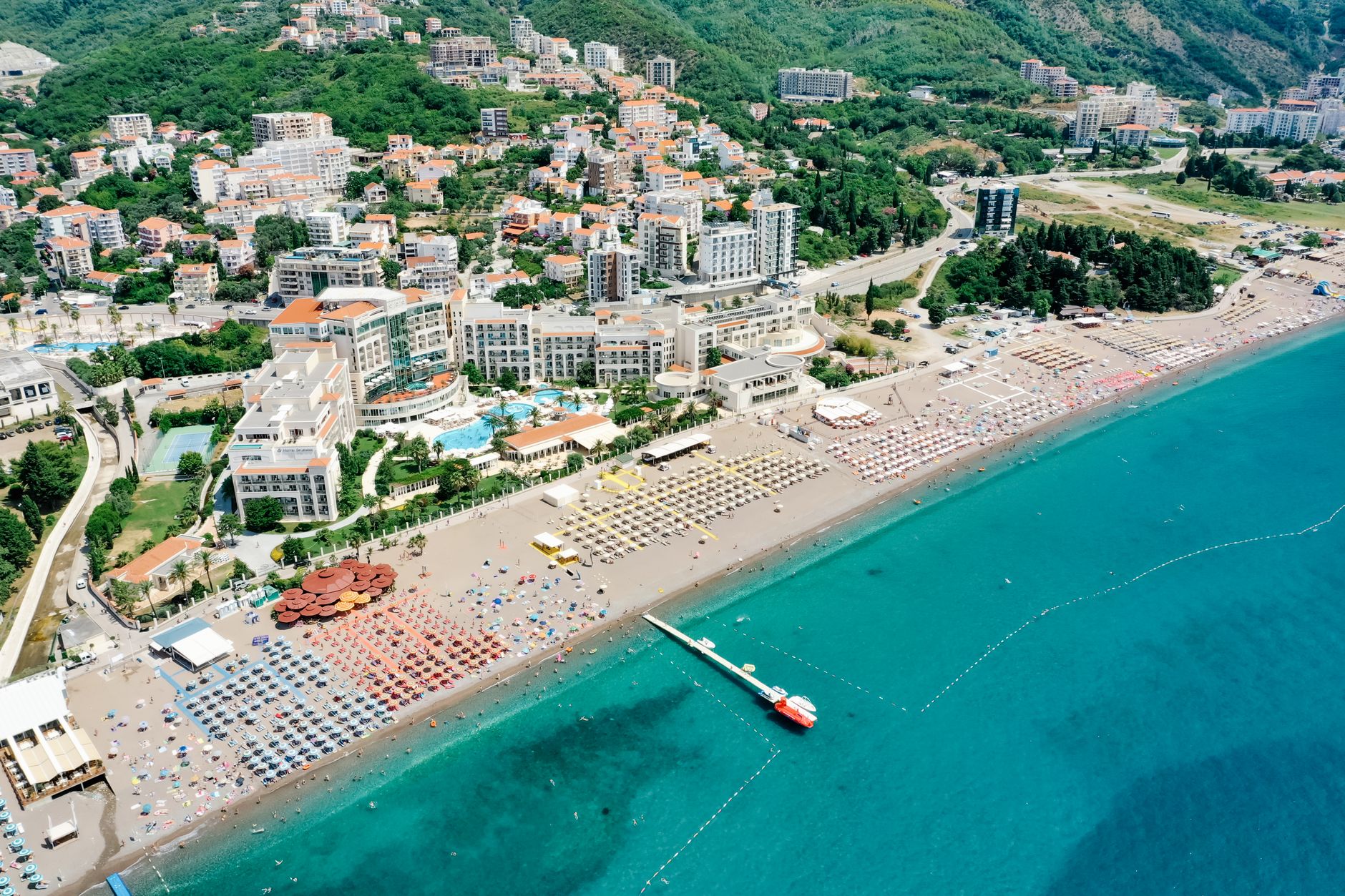 Aerial view of a long resort beach on the Budva riviera, ranks of sunbeds and umbrellas, hotels at the waterline and apartment blocks climbing the hill behind