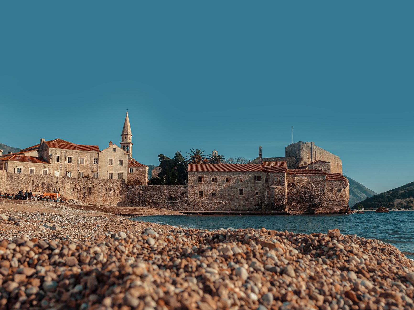 Budva's old town walls and the campanile of St John's seen across a beach of coarse rounded pebbles in low sun