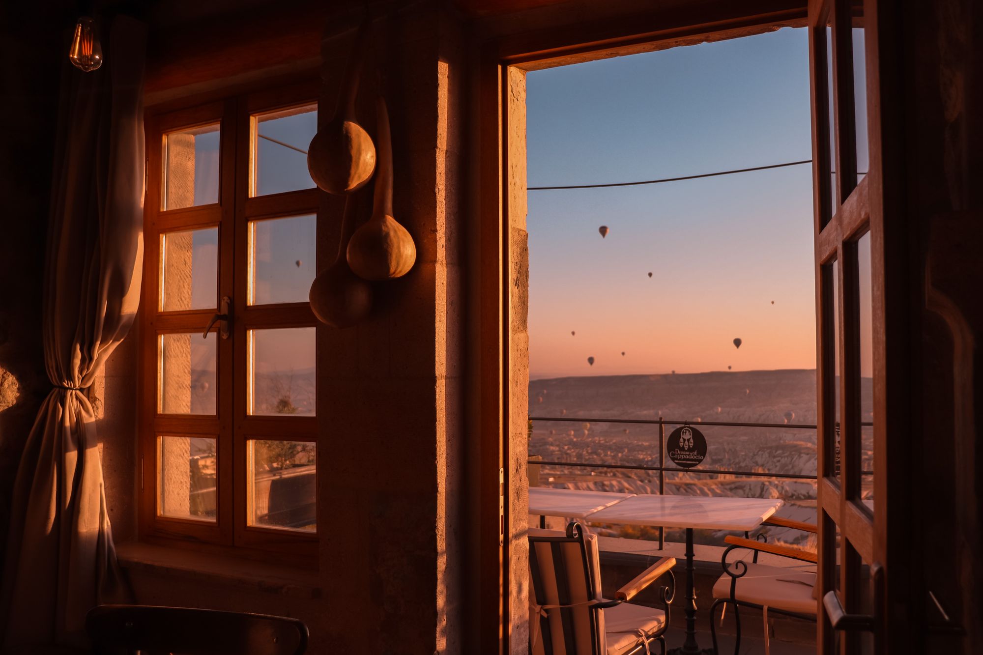 A cave hotel window in Cappadocia opening onto a terrace, balloons dotting the dawn sky outside