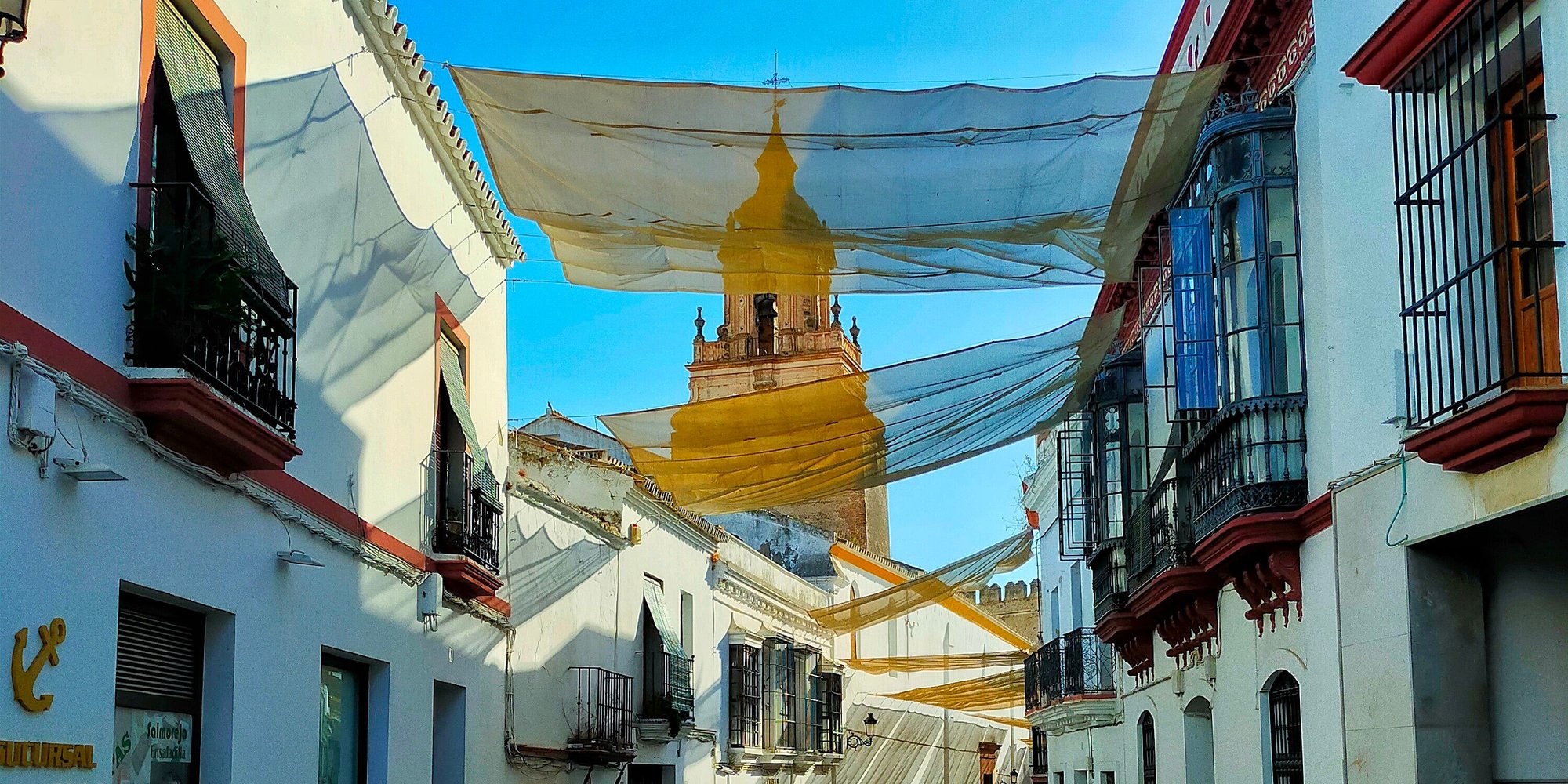 Summer awnings strung across a whitewashed lane in Carmona's old town, with a baroque bell tower and a crenellated wall at the end of the street