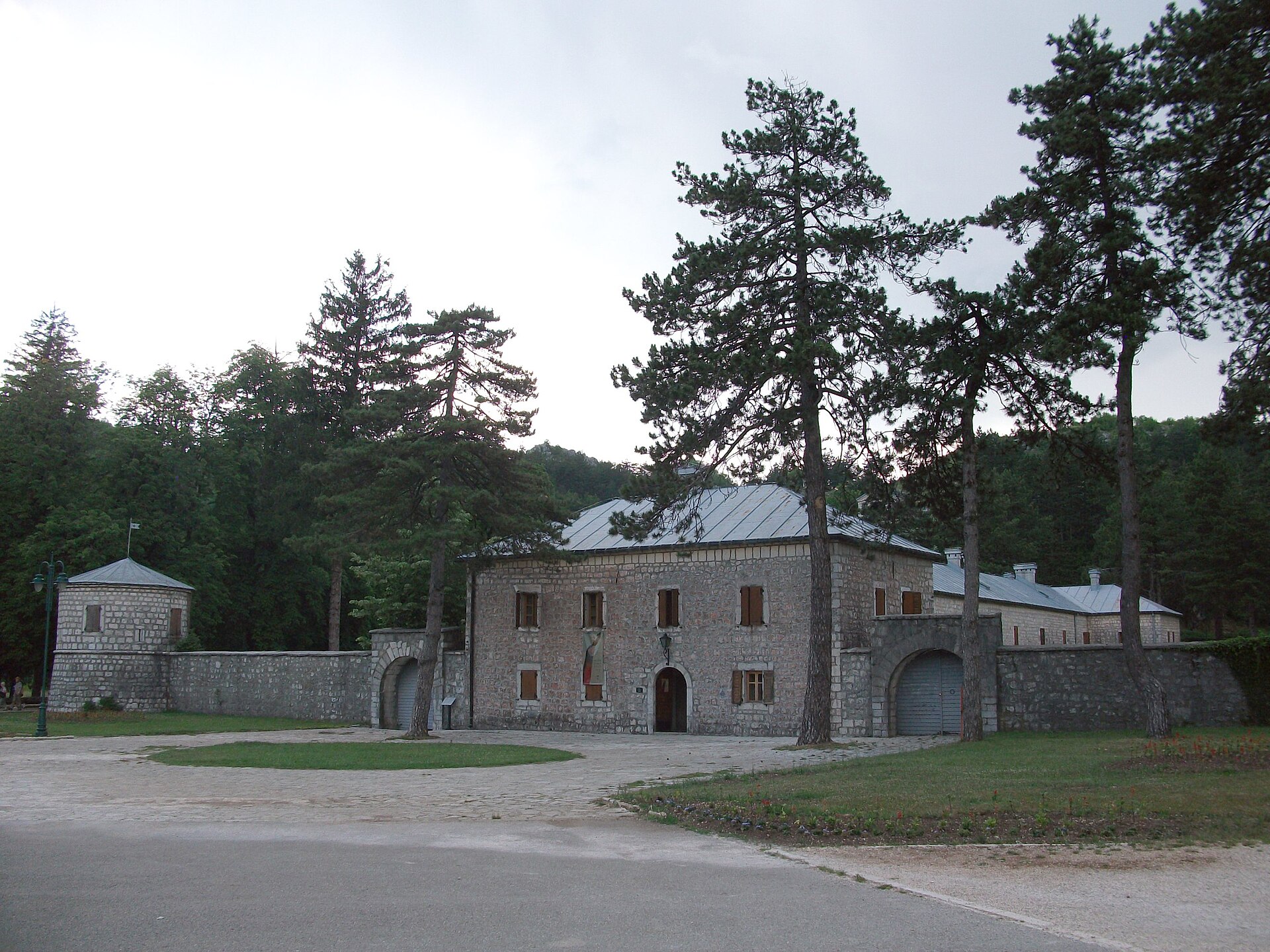 Biljarda in Cetinje, a two-storey stone building with a round corner tower and an enclosing wall pierced by arched gates, standing among tall pines