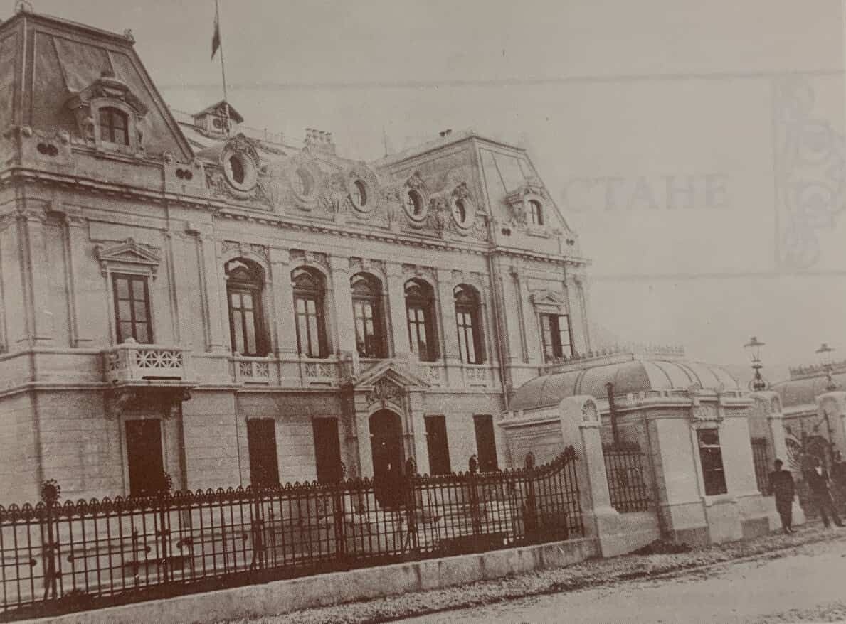 A sepia photograph from the early 1900s of the Russian legation in Cetinje, a heavy mansion with a mansard roof standing behind iron railings, with a figure at the gate
