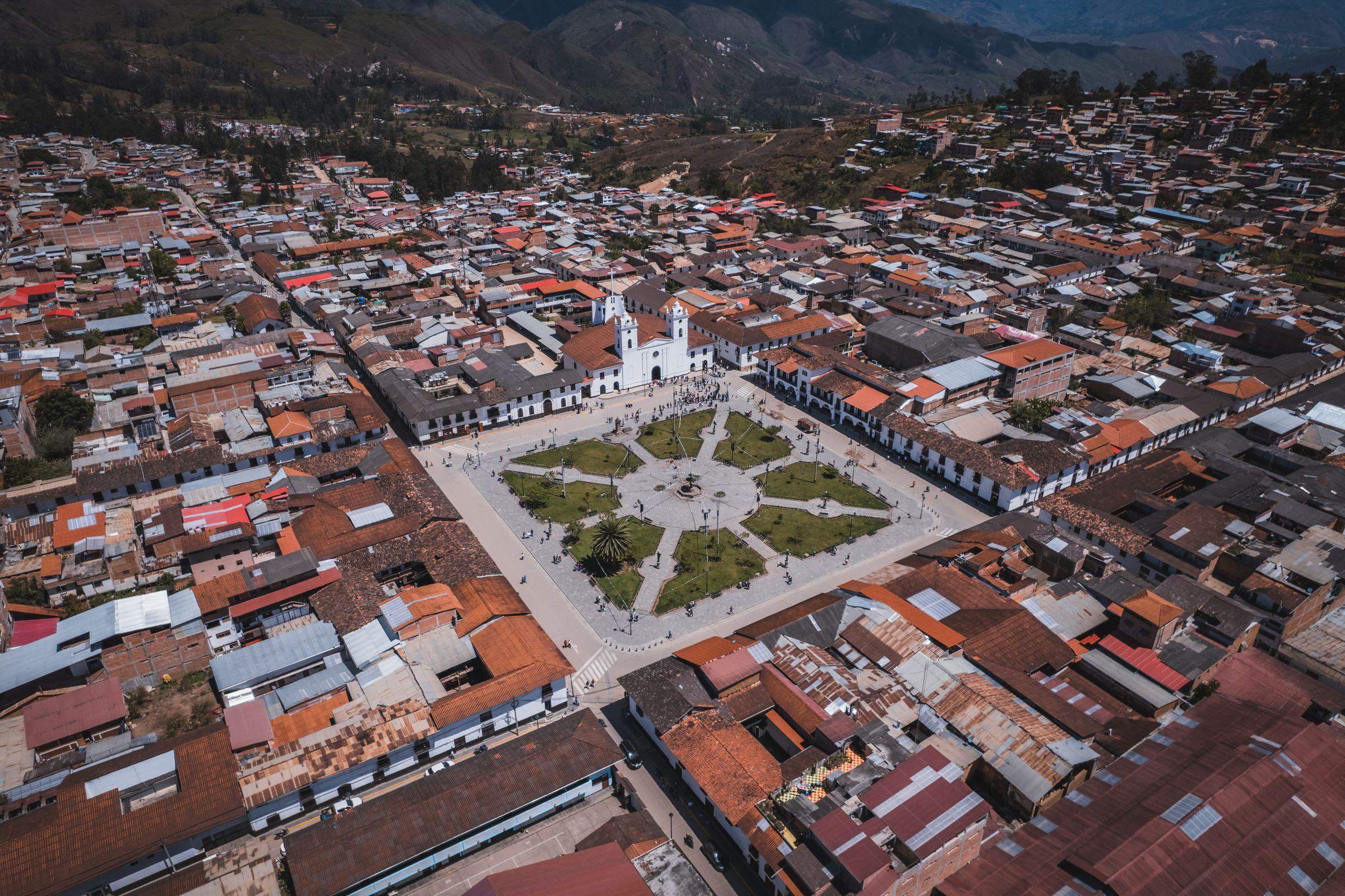 An aerial photograph of the Plaza de Armas in Chachapoyas, its gardens and the white cathedral, surrounded by red-tiled and tin roofs, with green hills behind