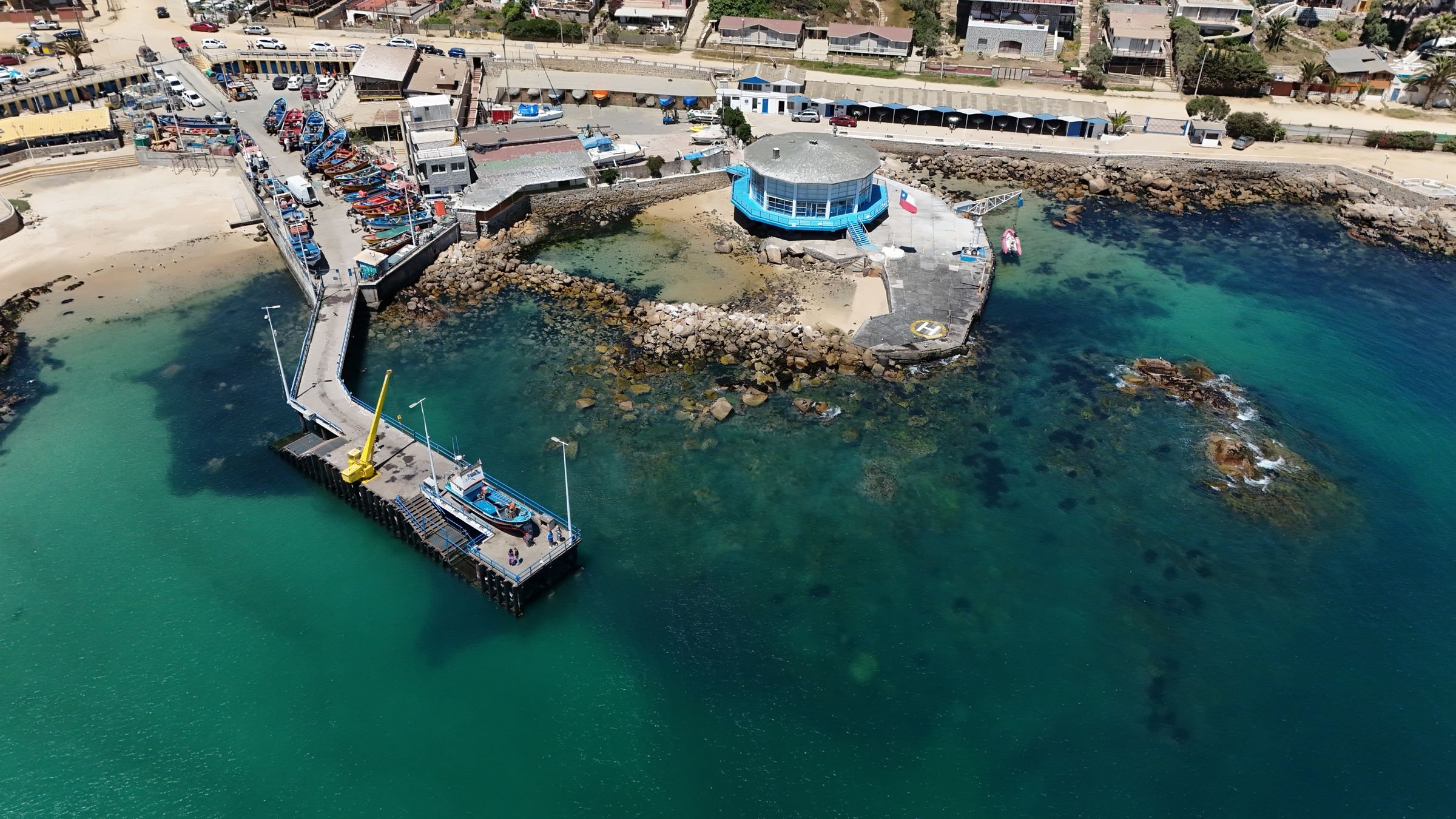 Algarrobo pier from the air, fishing boats moored in clear green water beside rocks and the town waterfront