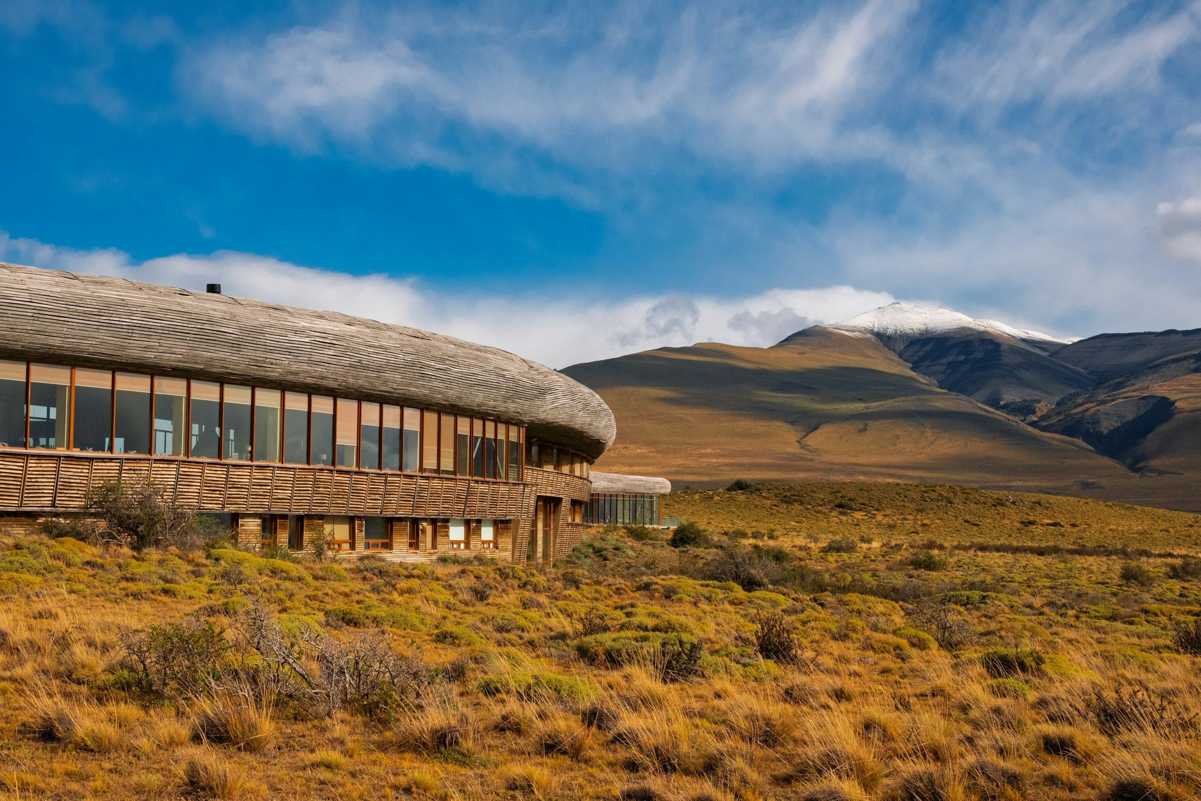 A long low hotel of curved timber set into the Patagonian steppe under a wide sky, with hills behind it
