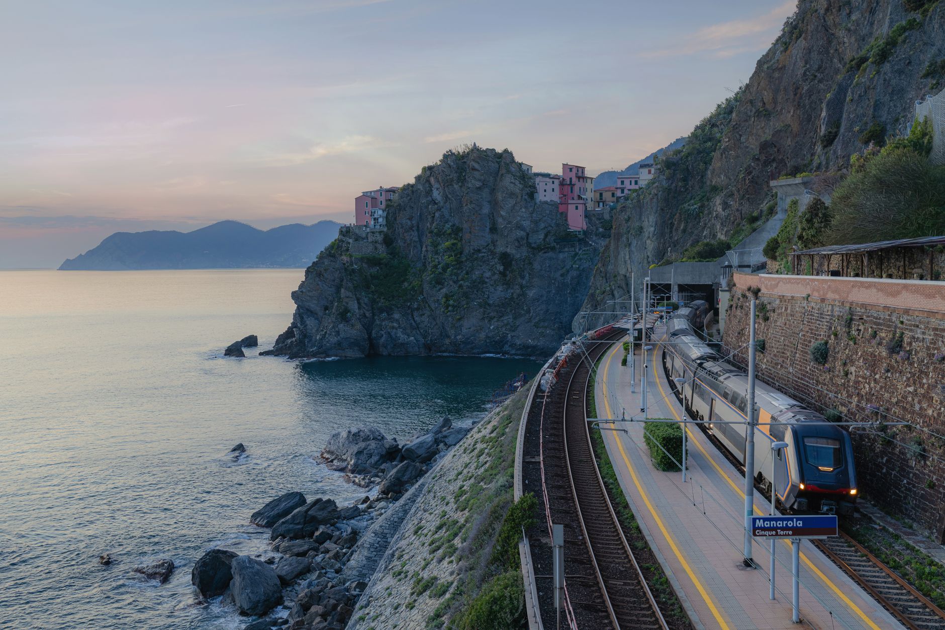 A regional train at a cliffside station in the Cinque Terre beside a turquoise bay and a tunnel mouth