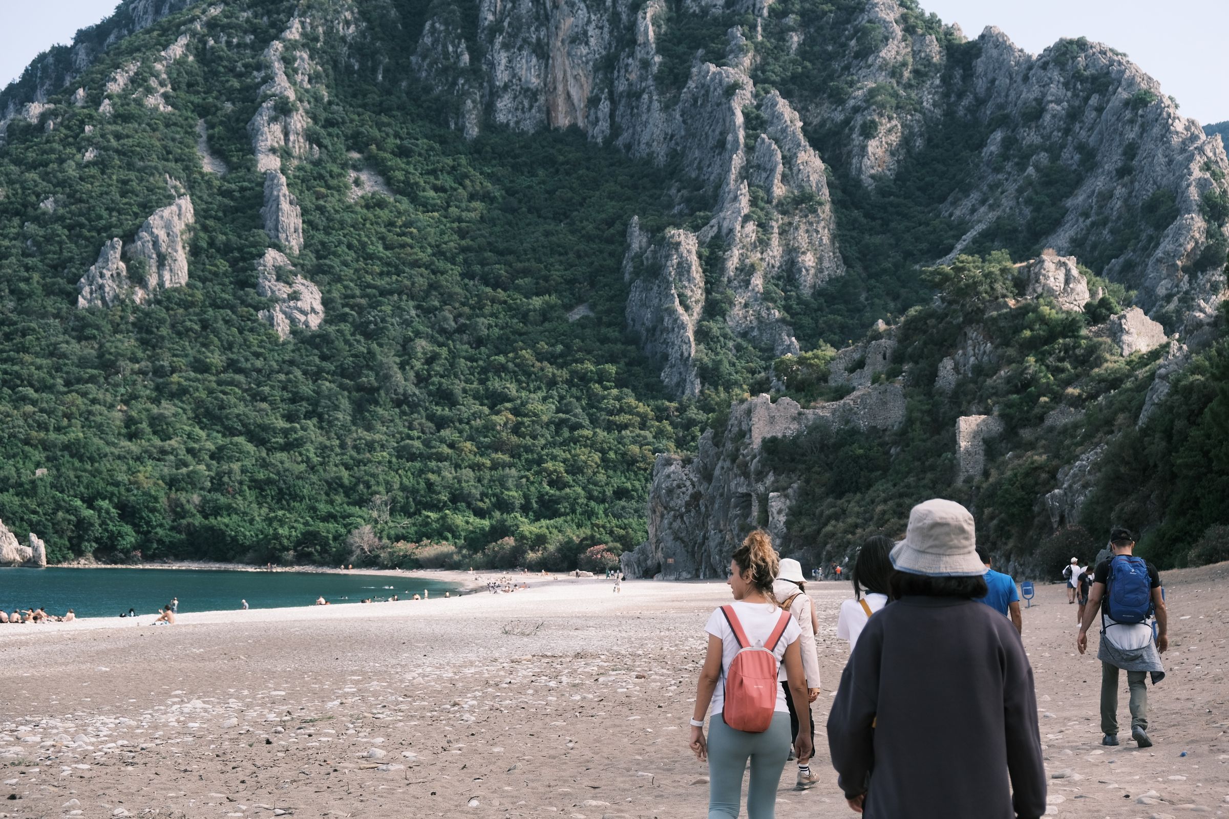 Walkers on the pebble beach at Olympos, below forested limestone cliffs with old stone walls above the shore