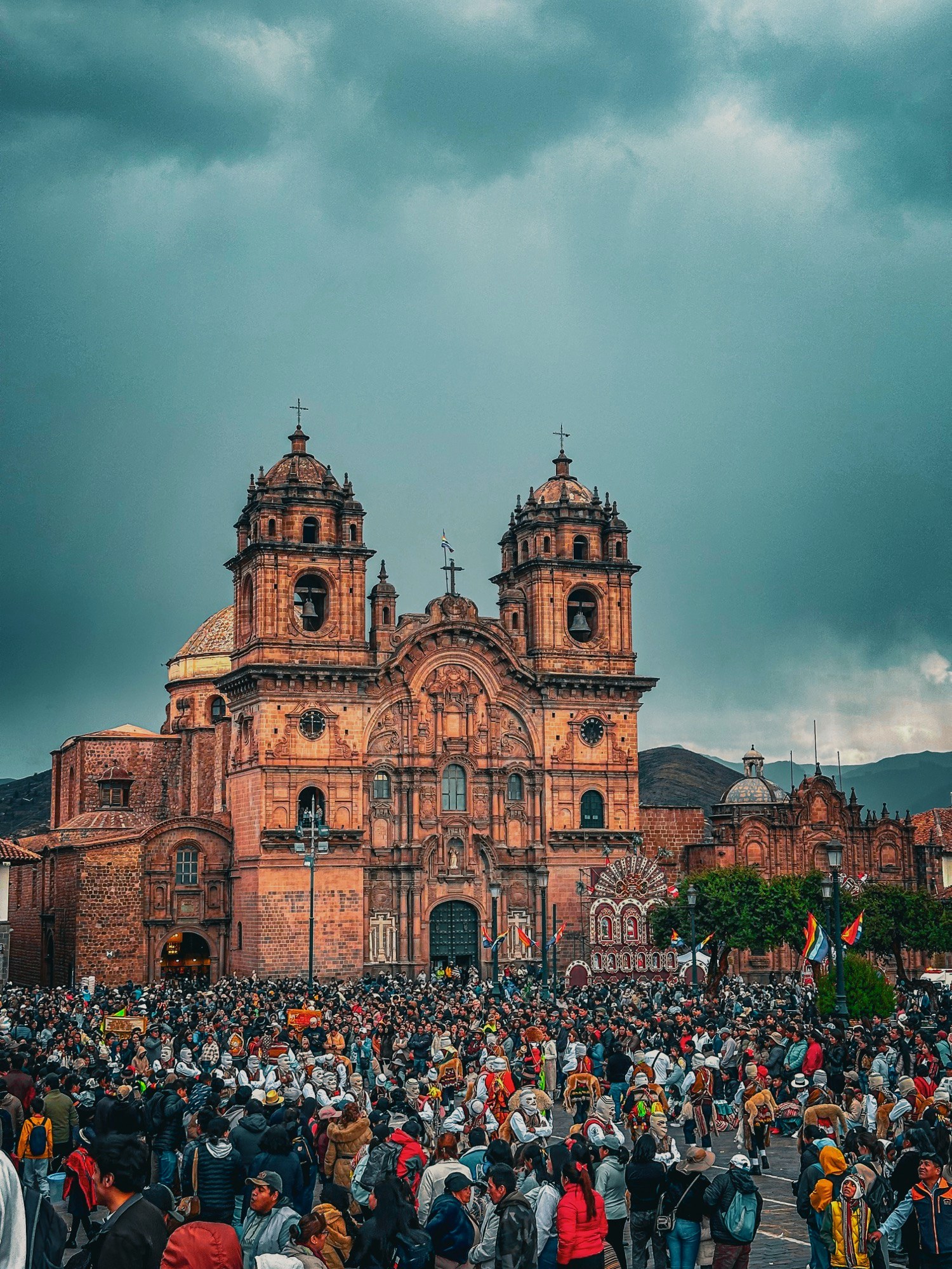 The baroque facade of La Compañía de Jesús church in Cusco rising over the festival crowd, dancers in white masks below
