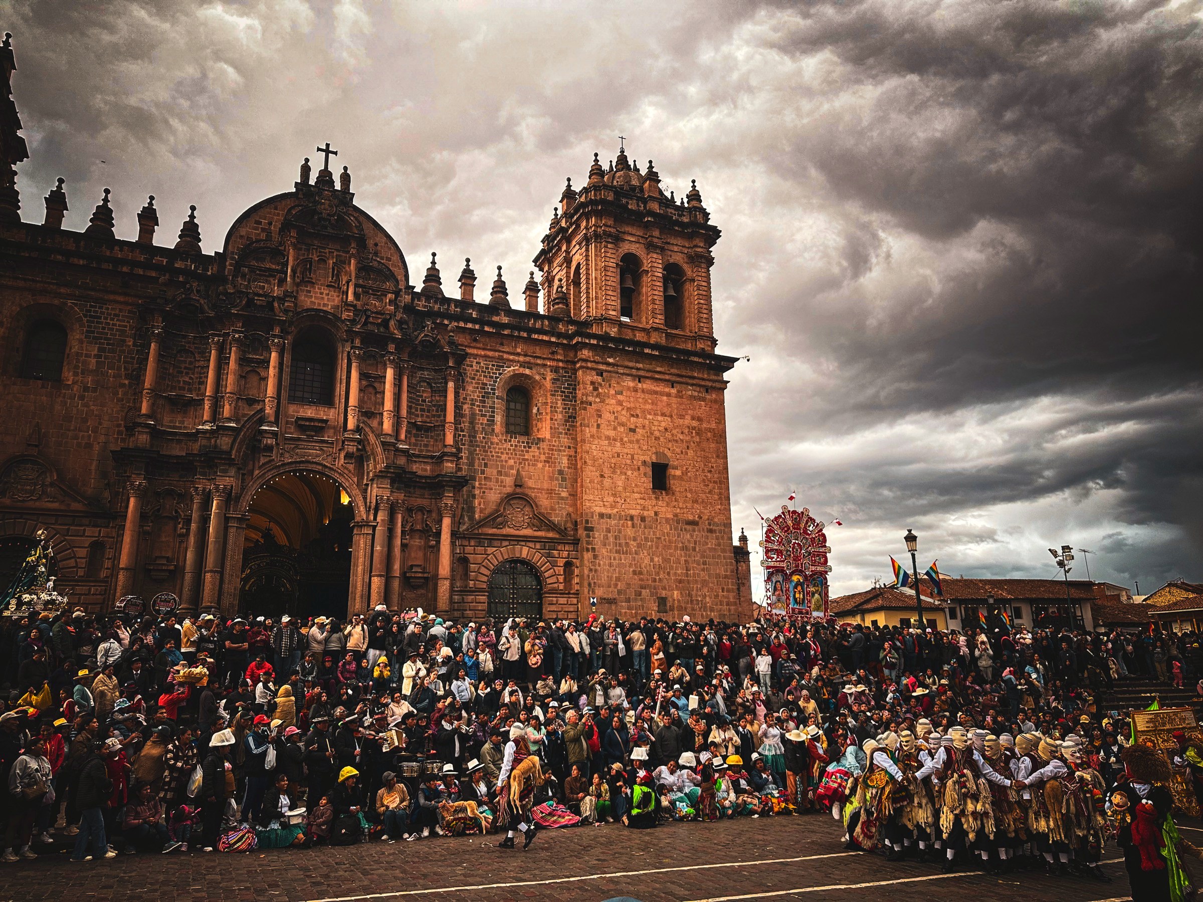 A huge crowd in front of Cusco cathedral under dramatic storm clouds, masked dancers gathered to one side waiting for the procession