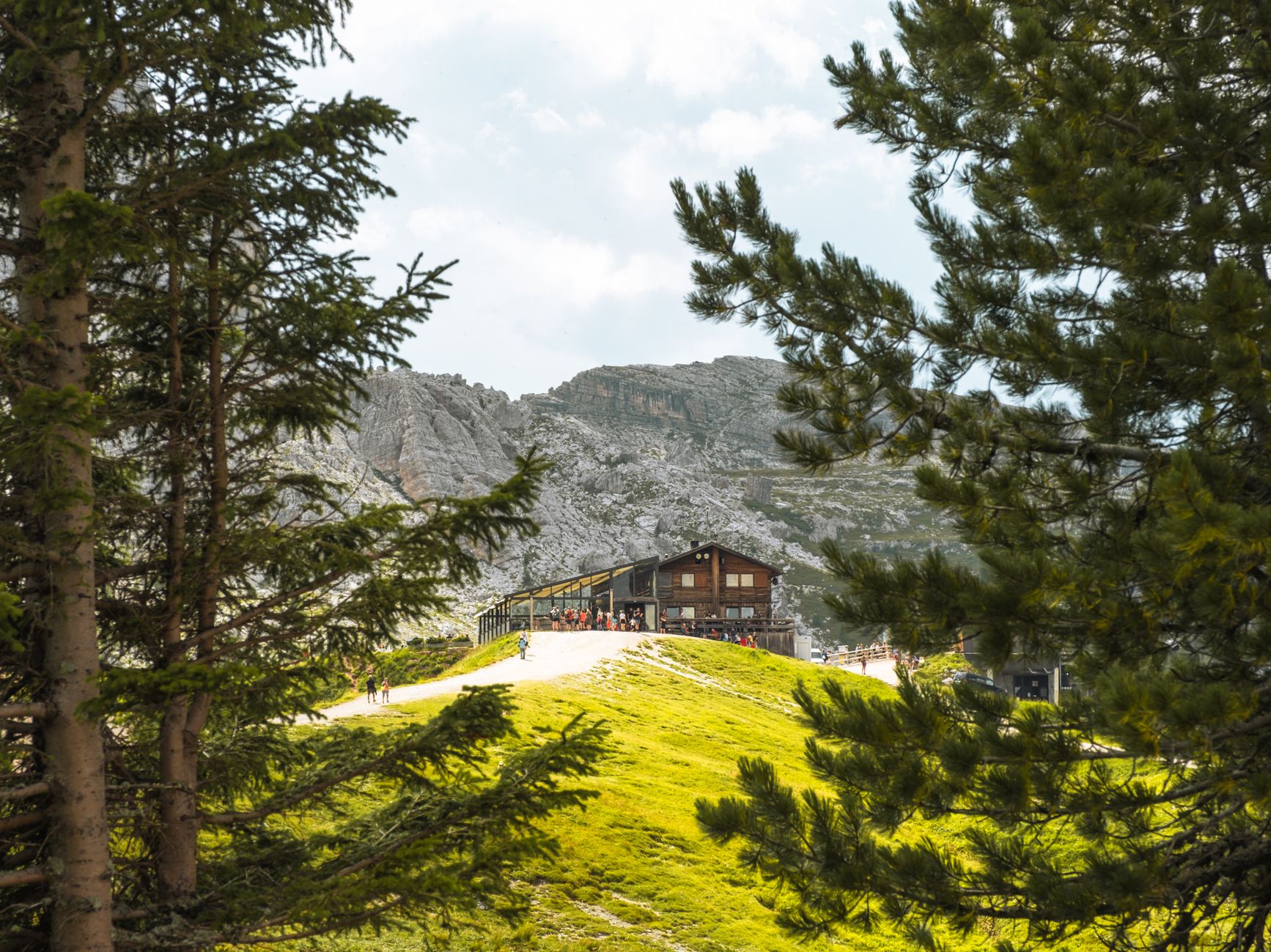 Alpine refuge on a green meadow shelf framed by pines below Dolomite walls