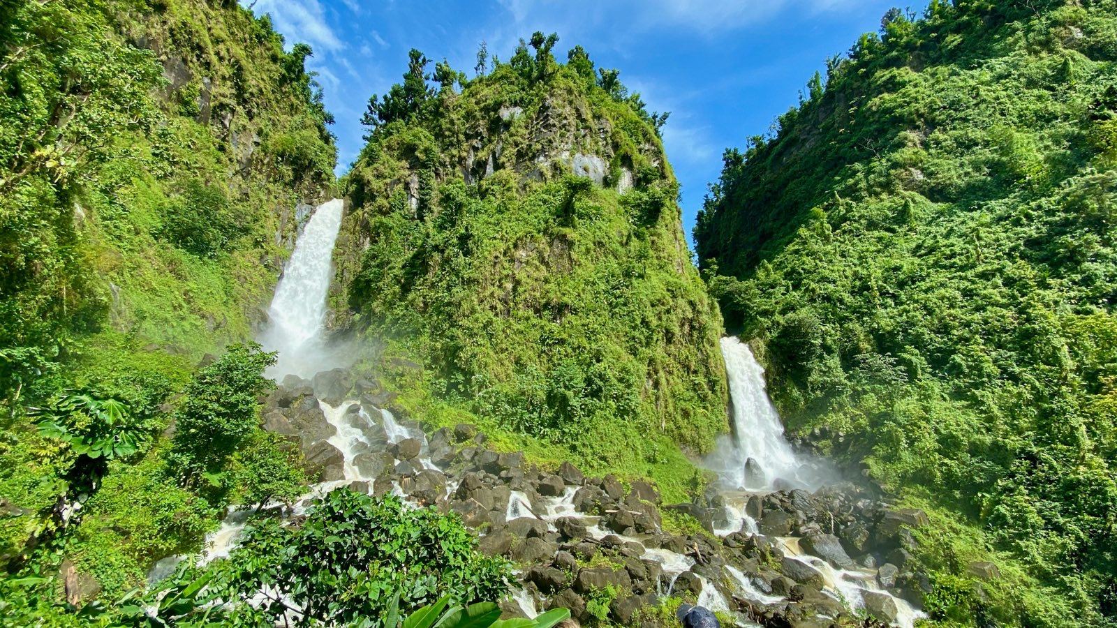 The two waterfalls of Trafalgar Falls dropping side by side down a green cliff onto black boulders under a blue sky